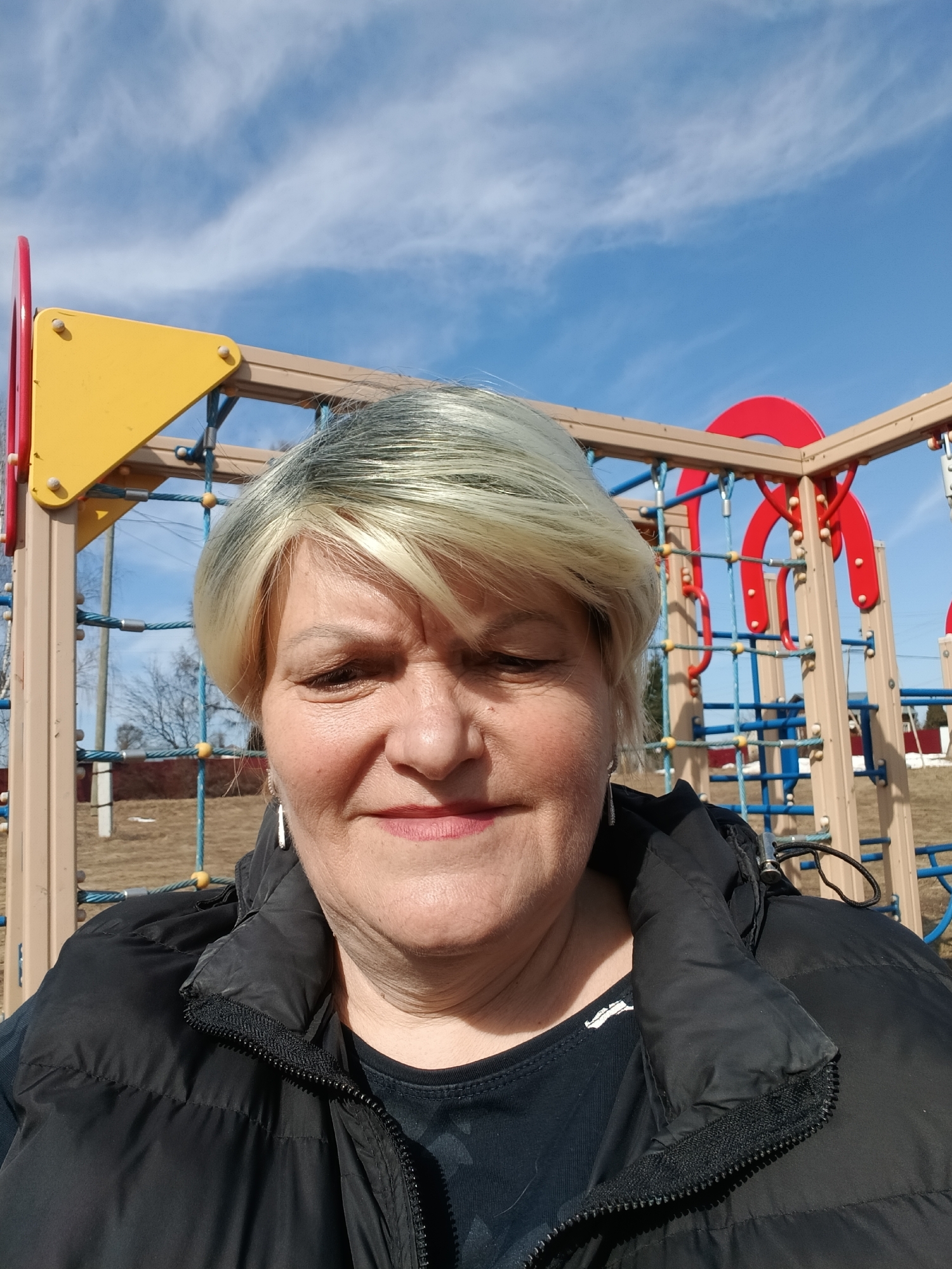 A selfie of a woman with short, blonde hair. She is wearing a black puffer jacket over a dark shirt. Behind her is a playground structure with yellow, red, and beige elements. The sky is blue with wispy white clouds. There is grass in the background, along with a couple of trees. The location is Красный Маяк, Russia.