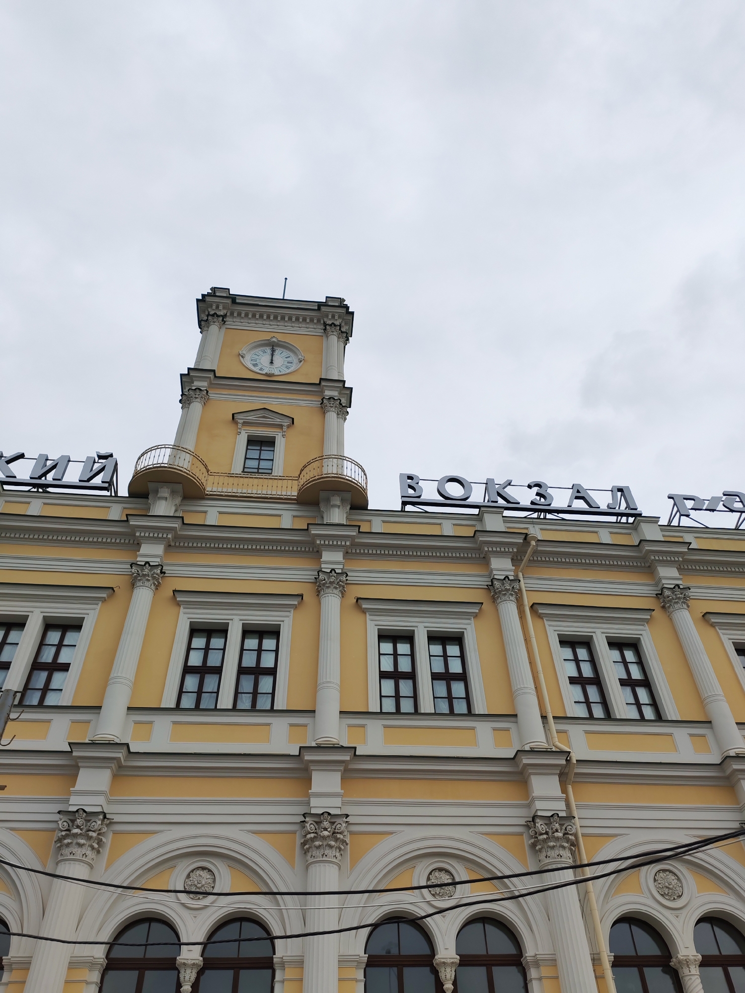The image presents a low-angle, upward view of a grand, ornate building under an overcast sky. The structure is predominantly light yellow with extensive white architectural detailing.
A prominent square clock tower rises centrally, featuring a circular clock face that indicates approximately 12:00. Below the clock, two small, curved balconies with decorative railings adorn the tower. The building's visible facade includes multiple levels of windows; the upper ones are rectangular with dark frames, while the lower level features arched windows, also with dark frames. The facade is adorned with white fluted columns and pilasters, topped with intricate Corinthian-style capitals and other decorative elements.
Large, dark, three-dimensional letters are mounted on the roofline. On the left, partially visible, are the Russian characters "КИЙ". On the right, the word "ВОКЗАЛ" (VOKZAL, meaning "station") is clearly spelled out in Russian, followed by "РА".
The sky is uniformly light grey and cloudy, indicating overcast weather. No people or specific activities are visible in the static architectural shot.