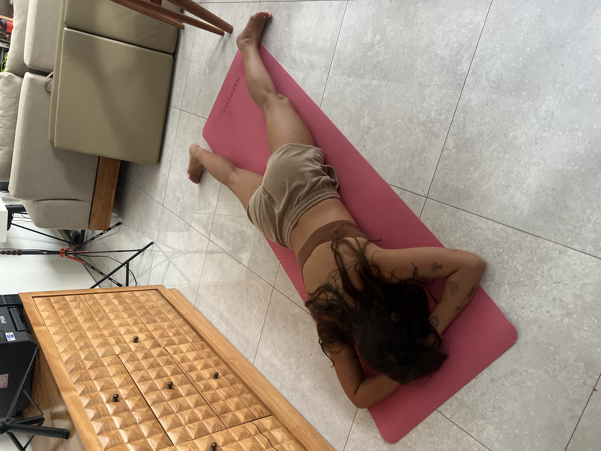 A woman is lying prone on a pink yoga mat on a tiled floor in what appears to be an indoor setting. She is wearing a brown sports bra and shorts. Her long, dark hair is spread around her head on the mat. Her arms are bent, with her hands near her head. The yoga mat has the word "HAPPYFIT" printed on it.
A light-colored sofa sits on the left side of the image, near the top. Next to it, a wooden table is visible, its surface featuring a patterned design.
A black tripod stands near the wooden table, and there is a printer visible on the left side of the image, behind the wooden table. The background consists of the tiled floor.
This image does not provide sufficient details to accurately identify a specific location in Mengwi, Indonesia.