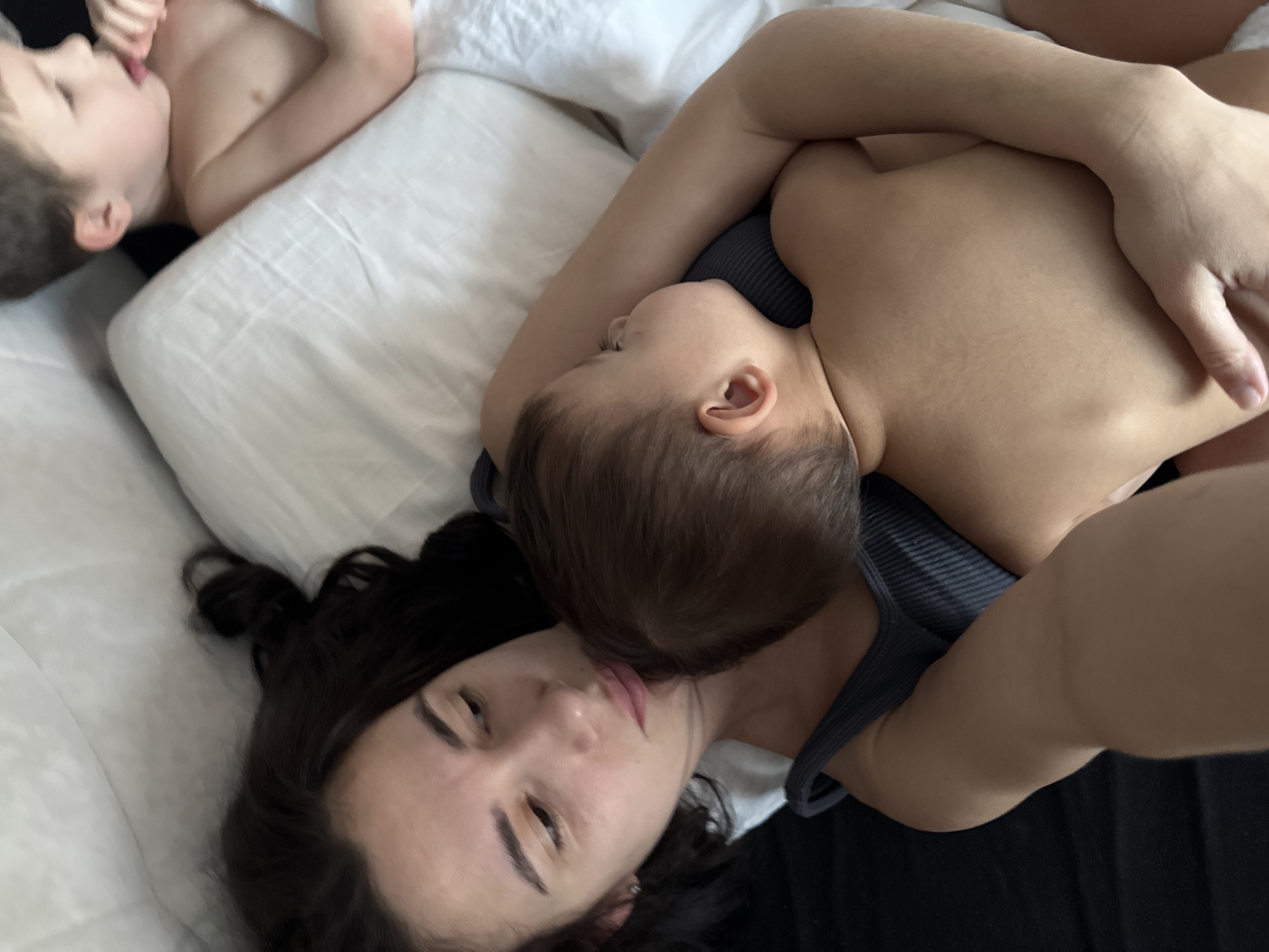 The image, taken in the city of Chaikovsky, Russia, presents an overhead view of a woman and two children in a bed. The woman, positioned on the left and central, has dark hair and is wearing a dark grey ribbed tank top. She lies on a dark sheet, looking to her right (viewer's left) with a neutral expression. Her left arm extends towards the bottom of the frame, presumably holding the camera, while her right arm wraps around a bare-chested toddler. This younger child, with light brown hair, is cradled against the woman's chest, facing away from the camera. In the upper right portion of the frame, an older bare-chested boy with light brown hair is visible, lying on white bedding. He is depicted in profile, looking to his left, with his hand near his mouth. The bed features white pillows and sheets, with a dark sheet covering the area beneath the woman and the left side of the frame. All three individuals appear to be resting or relaxing.