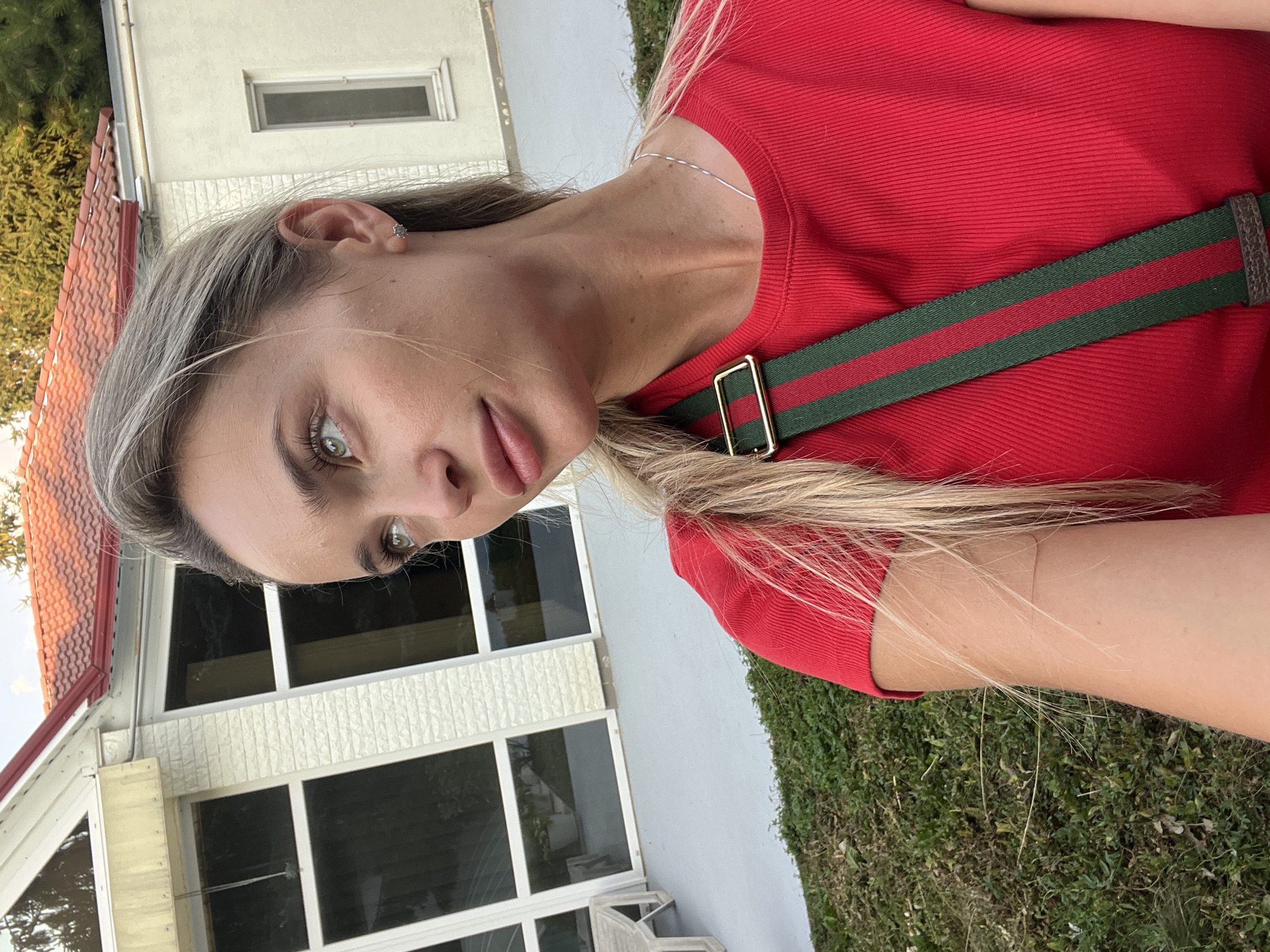 An outdoor, medium shot captures a blonde woman looking to her right, with her head slightly tilted. She is wearing a red, ribbed t-shirt with a green and red striped strap of a bag crossing her chest. Her hair is pulled back into a ponytail, with some strands falling forward. She is wearing a small, stud earring on her right ear and a thin silver necklace. The background features a white, brick-like textured building with large windows and a red tiled roof. Green grass and some trees are visible in the foreground and background, suggesting a suburban or rural setting. The scene appears to be a daytime photograph, with soft lighting. There is no information available to confirm the location in the city of Fontanka, Ukraine.
