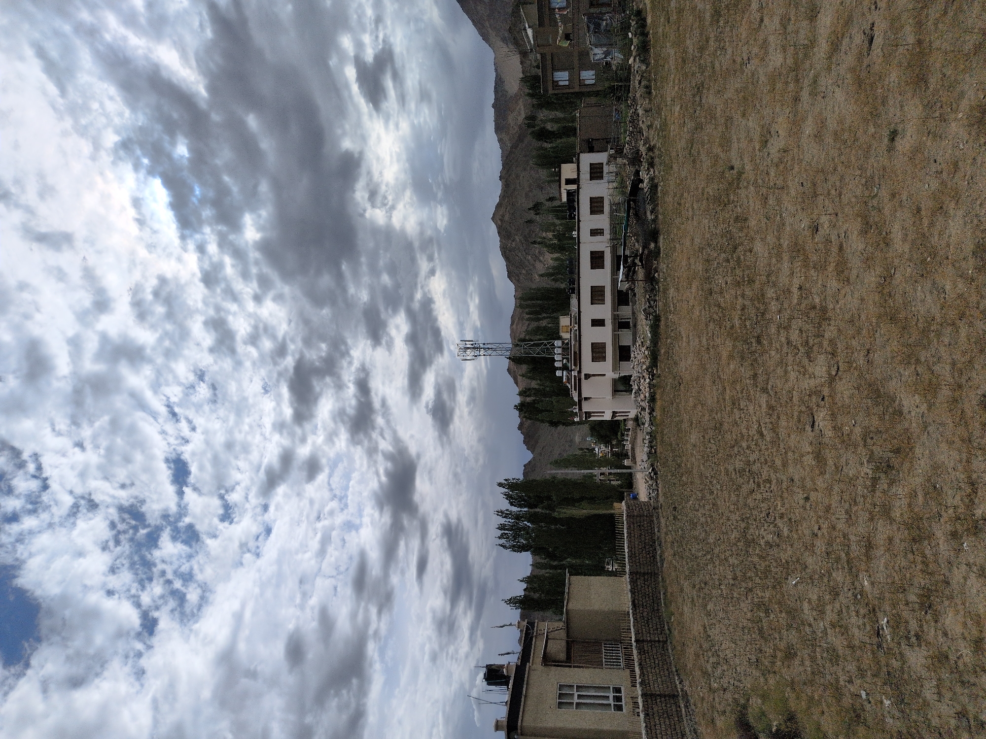 The image shows a wide shot of a semi-arid landscape with buildings and a cellular tower. The location is Leh, India. In the foreground, there is a dry, grassy field with sparse vegetation. To the left, a two-story building with a light-colored facade and windows is visible, behind a stone wall. Further to the right, a long, white building with multiple windows and a flat roof is situated. A tall, metal cellular tower stands in front of this building. Behind the buildings, a line of dark green trees is visible, and beyond that, a range of brown, rocky mountains stretches across the horizon. The sky is cloudy, with dramatic grey and white formations. There are no people visible in the image.