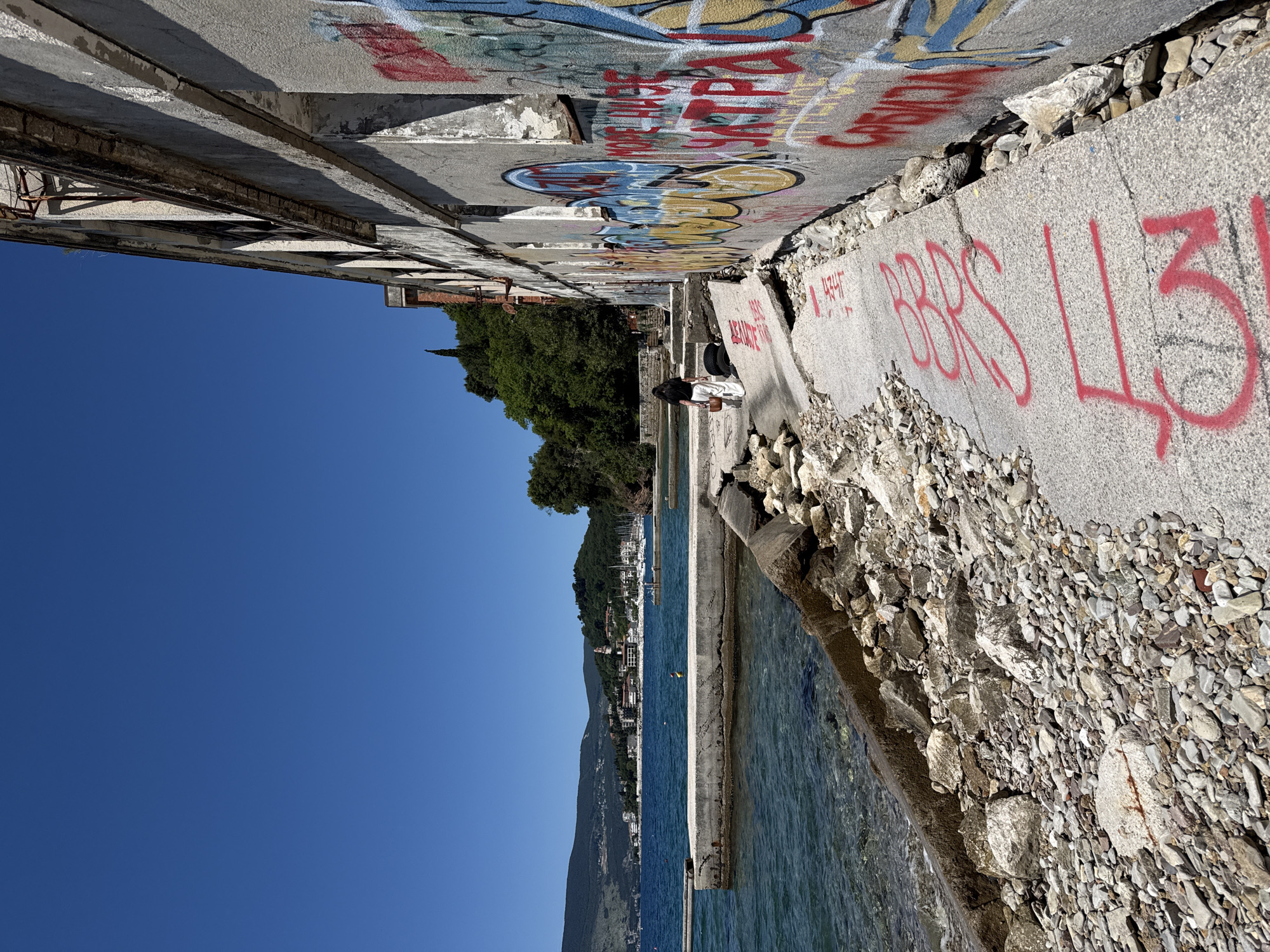 The image shows a waterfront scene in Sasovići, Montenegro. A dilapidated concrete structure with graffiti lines the water's edge. A woman in white pants and a dark top stands near the structure. The clear blue sea stretches out to a distant shoreline with buildings and green hills. The sky is a clear, vibrant blue. The concrete structure is in disrepair, with crumbling edges and exposed rocks. Red spray paint covers parts of the concrete.