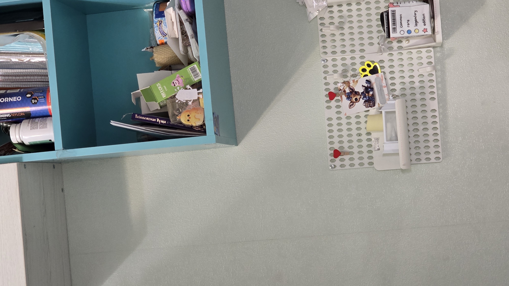 The image shows a close-up view of a light blue shelf unit containing various craft supplies. The shelf contains a bottle of white pills, a spool of orange nylon thread, a box of "BRICKS" craft items, and other small objects. Below the shelf, a pegboard is mounted on a pale green wall, decorated with small heart-shaped clips and a collection of charms and cards. One card displays different rarity levels with corresponding colored circles, and another features a cartoon giraffe. The pegboard also holds a small shelf with a roll of tape and a white rectangular object. The scene appears to be indoors, likely a home or craft room. There are no people visible in the image.