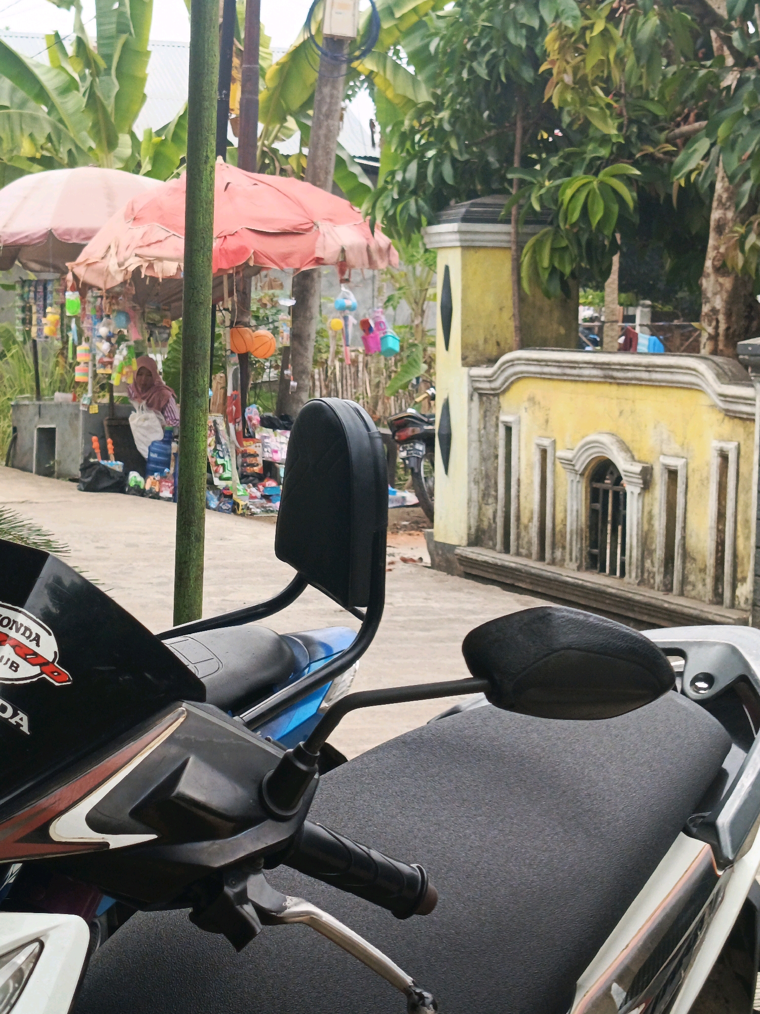 This image depicts a roadside scene in Lubuklinggau, Indonesia, likely a commercial area given the presence of small stalls and merchandise. In the foreground, two motorbikes are parked, with the closest one showing a black seat and handlebars, and a partial view of its front section with "HONDA" branding. Behind the motorbikes, a woman wearing a hijab is visible, standing near a stall displaying various items, possibly toys and household goods, under pink umbrellas. The background features a yellow wall with decorative features, lush green trees, and banana plants. The overall impression is of a vibrant, everyday street scene in a tropical setting.