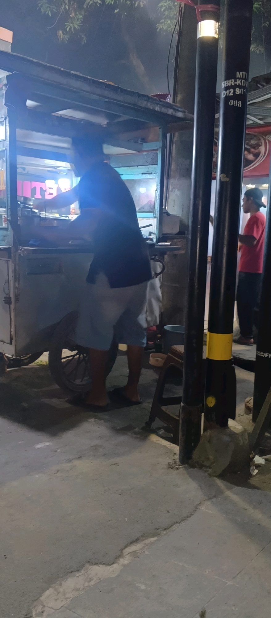 The image depicts a street scene at night in Kedungdawa, Indonesia, likely near a food stall. In the foreground, a person wearing a dark blue shirt and light-colored shorts is seen from the waist down, standing next to a food cart. The cart appears to be made of metal and wood, with a canopy. Behind the cart, a metal pole with reflective bands is visible, bearing markings that read "MTI", "SBR-KDV", "D12 S01", and "088". Further back, another person wearing a red shirt and a dark cap is standing near a building. The ground is paved and appears somewhat uneven. Trees with foliage are visible in the background, and artificial lighting from the street or the food stall illuminates the scene.