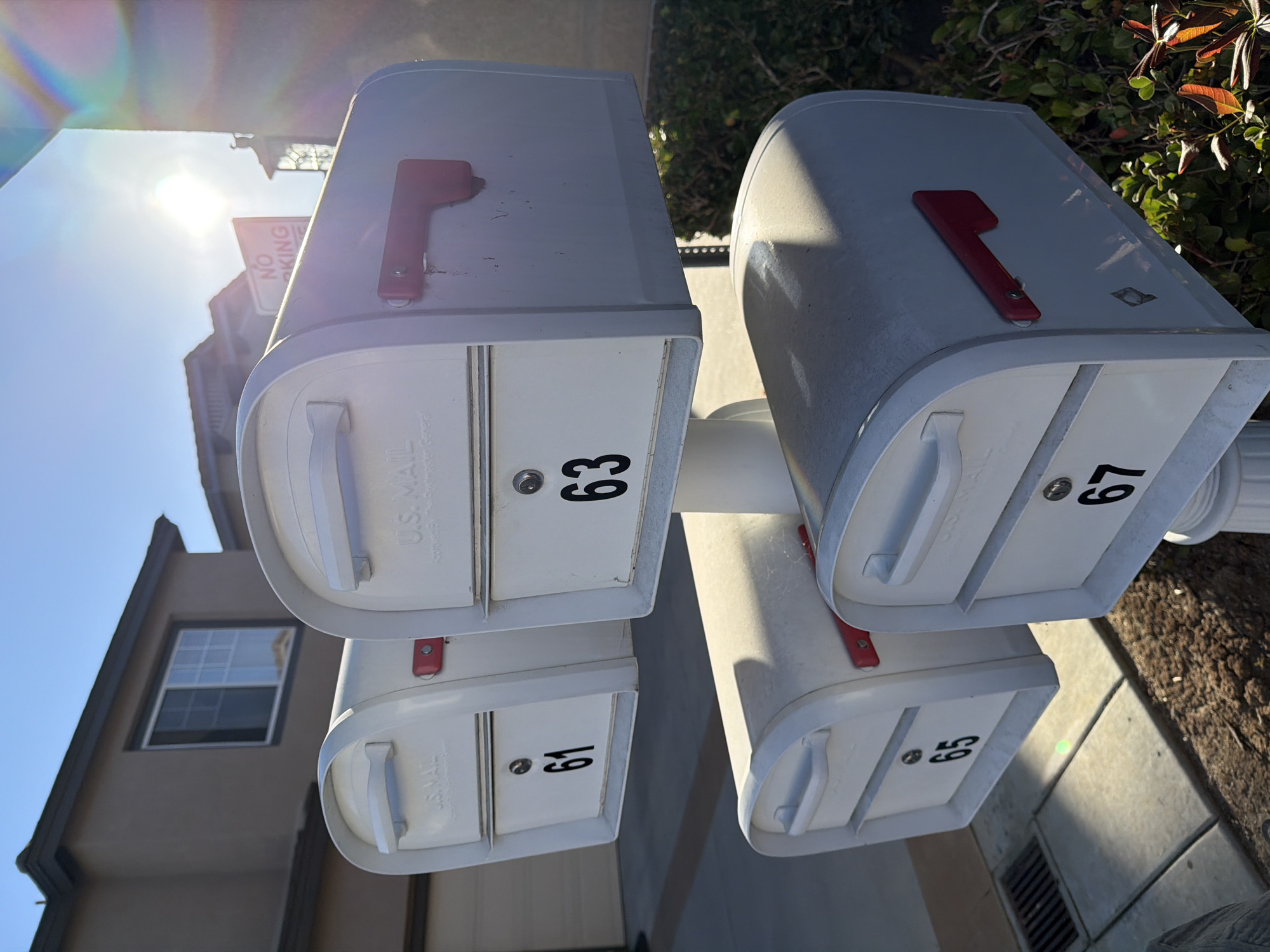 A row of four white U.S. Mail mailboxes, numbered 61, 63, 65, and 67, are arranged in a two-by-two configuration on a single post in Aliso Viejo, California. The mailboxes are made of metal and have red flag latches. They are positioned outdoors in front of a beige residential building with a dark roof and a window. A "NO PARKING" sign is visible in the background, partially obscured by the sun's glare. The scene is illuminated by bright sunlight, casting shadows and creating lens flare. The mailboxes are set against a backdrop of green bushes and a paved walkway. The image appears to be taken during daylight hours.