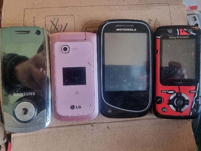 The scene is located in Balgarska Poliana, Bulgaria. Four mobile phones are displayed horizontally from left to right on a flat, light brown corrugated cardboard surface. The cardboard shows faint black printed text and symbols.
From left to right:
1. A silver Samsung mobile phone, possibly a slider or candy bar form factor, with a reflective screen displaying a partial reflection of a hand or arm in dark clothing. A circular, silver control module is present at the bottom front.
2. A closed, pink LG flip phone. The exterior shows a small, dark display panel and a visible camera lens. The surface exhibits multiple scratches and wear marks.
3. A black Motorola smartphone with a dark, off-screen that appears dusty and smudged. The "MOTOROLA" logo is visible at the top, and a stylized 'home' icon is at the bottom.
4. A red and black Sony Ericsson F305 phone. Its screen is dark and dusty. The "Sony Ericsson" logo and "F305" model number are visible, along with a prominent circular control pad featuring gaming-style buttons in black and silver.
All phones appear to be powered off. No active interactions or individuals are directly visible in the frame, beyond the reflection on the leftmost device.