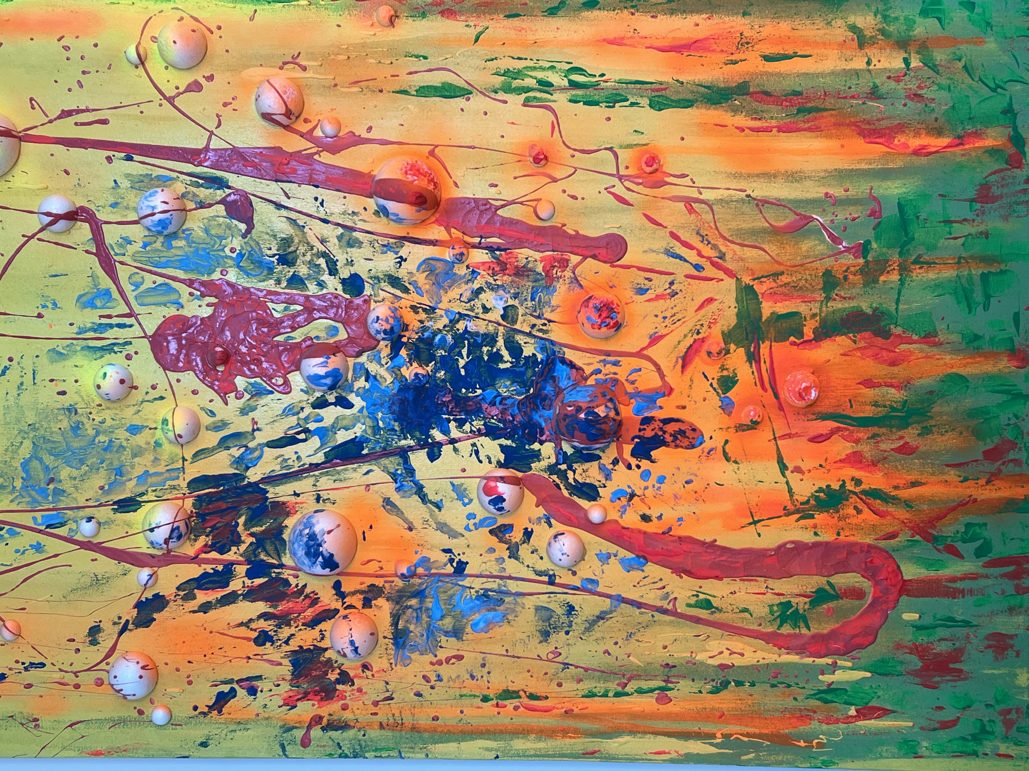 The image depicts an abstract painting with a vibrant and chaotic composition. The background is a blend of yellow, orange, and green, with visible brushstrokes creating a textured effect. Splatters and streaks of blue, red, and black paint are scattered across the canvas, adding to the energetic feel. Three-dimensional spheres of varying sizes are attached to the surface, some painted with designs resembling planets or abstract patterns. The artwork does not reveal any recognizable location or subject related to the city of Chaikovsky, Russia.