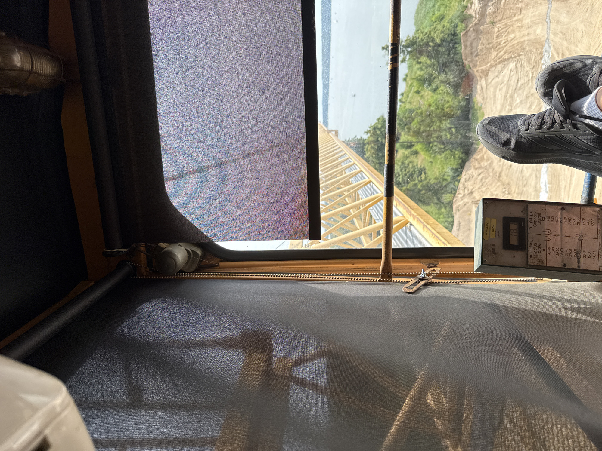The image is taken from the perspective of someone sitting in the cab of a crane, looking out a window. In the foreground, a person's black athletic shoes are resting on the window ledge or a similar surface. The shoes appear to be worn. Through the window, a yellow crane boom is visible extending over a body of water, likely Lake Mikołajskie, given the context of Mikołajki, Poland. Trees and sandy terrain are visible on the opposite shore. A control panel with some gauges and text in what appears to be Polish and German is also visible on the right side of the frame. The overall scene suggests a construction or industrial site near a body of water.