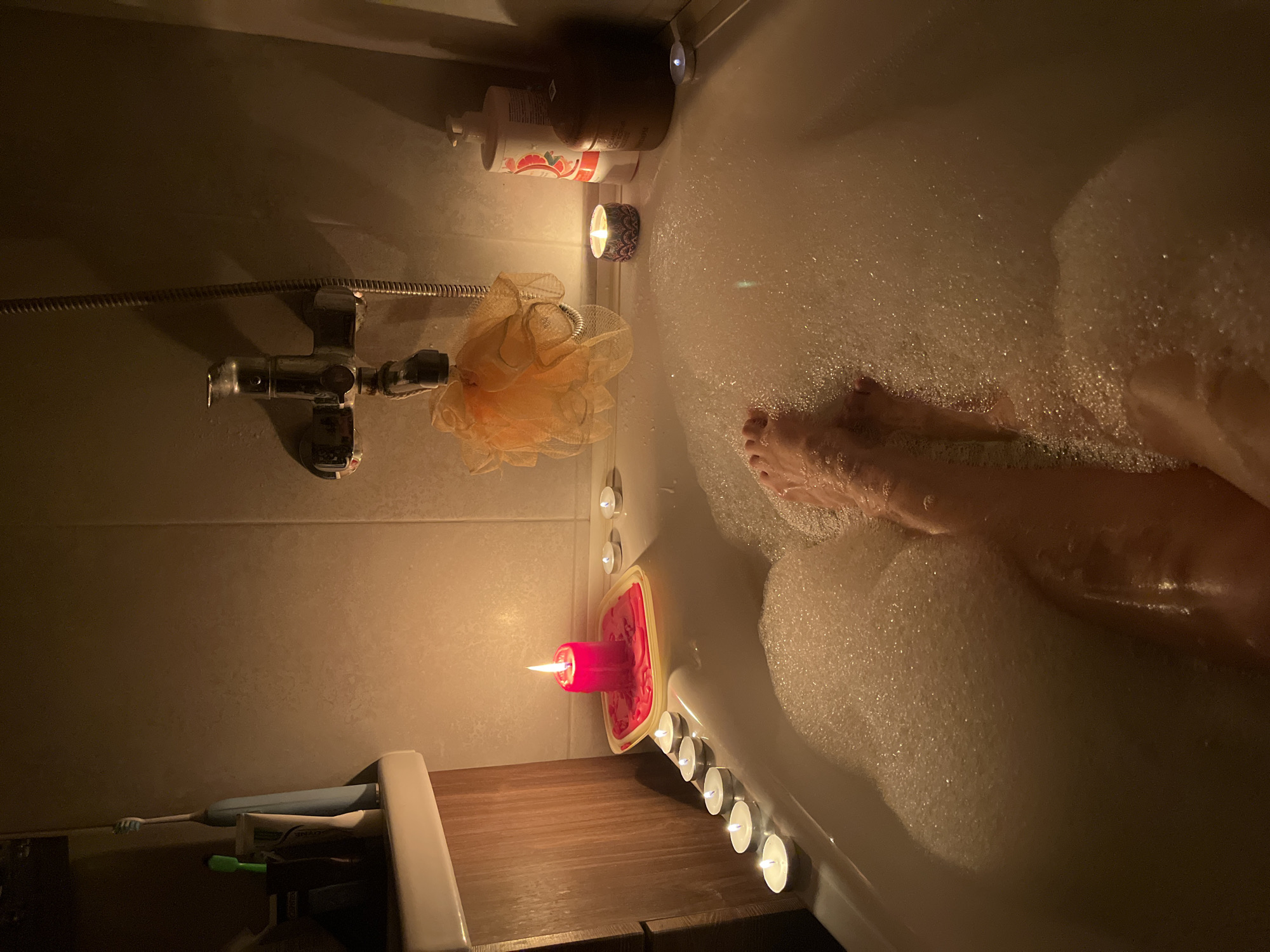 An overhead, medium shot captures a person's legs submerged in a bubble-filled bathtub, lit by the warm glow of numerous candles. The scene is set in a bathroom, possibly in Kazan, Russia, given the context. The bathtub is pristine white, filled with an abundance of white bubbles that rise to the surface. The person's feet and lower legs are visible, partially obscured by the foam.
Along the edge of the bathtub, several small tealight candles are arranged, casting gentle light. A larger, red pillar candle sits in a yellow dish on the left side, its flame dancing. Further along the edge, closer to the faucet and showerhead, are more tealight candles and a decorative candle in a patterned holder.
A bright orange loofah hangs from the showerhead, adding a splash of color to the otherwise subdued palette. To the right, a tall, white dispenser bottle with a red label and a wooden-lidded container are visible. The tiled walls of the bathroom are dimly lit, reflecting the candle glow. On the far left, a wooden cabinet or shelf holds two toothbrushes and a tube of toothpaste. The overall atmosphere is one of relaxation and tranquility, enhanced by the soft lighting and the presence of the candles.