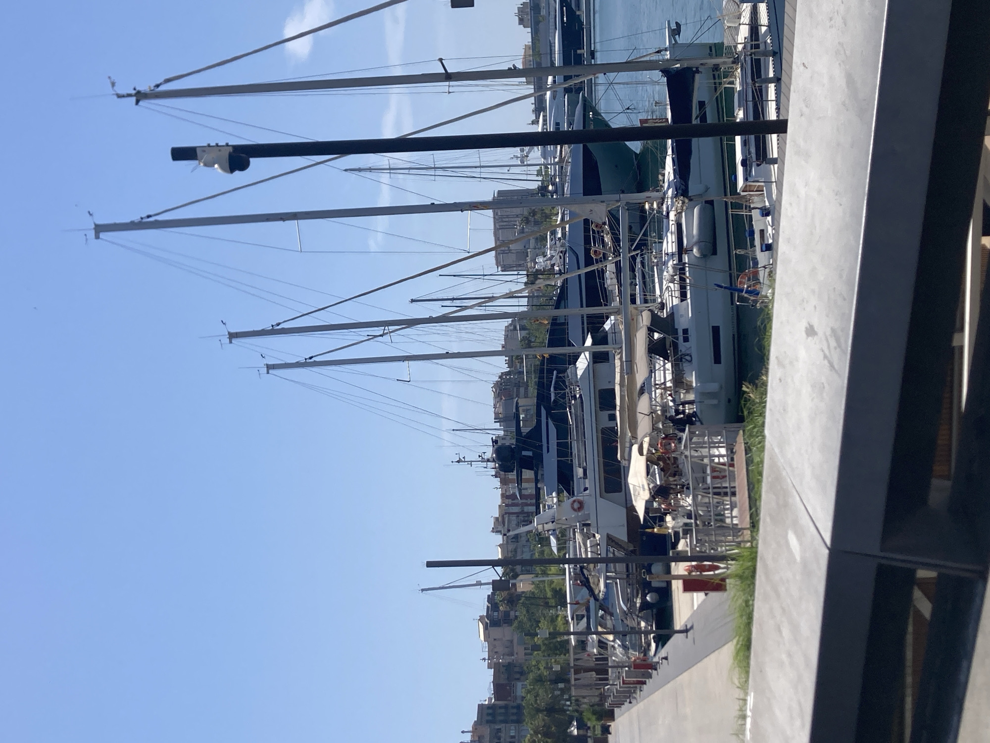The image depicts a harbor scene in Barcelona, Spain, on a clear day. A prominent feature of the image is a row of docked yachts and sailboats, their masts creating a vertical pattern against the bright blue sky. The yachts are of varying sizes and designs, with some appearing to be luxury vessels. In the background, buildings and greenery line the waterfront, suggesting an urban or semi-urban setting.
The perspective of the image is from a slightly elevated vantage point, looking across the docked boats towards the shore. A concrete walkway or pier occupies the foreground on the right side of the frame, with some vegetation growing along its edge. A black pole with a camera or sensor attached is visible in the middle ground, mounted on one of the boat masts.
The lighting suggests it is daytime, likely late morning or early afternoon given the brightness and clear sky. No specific activities or events are clearly discernible, other than the boats being docked. There are no visible people prominently featured, though some figures might be present on the decks of the boats, too small to identify. The overall mood is peaceful and serene, typical of a harbor on a calm day. No text is visible in the image.