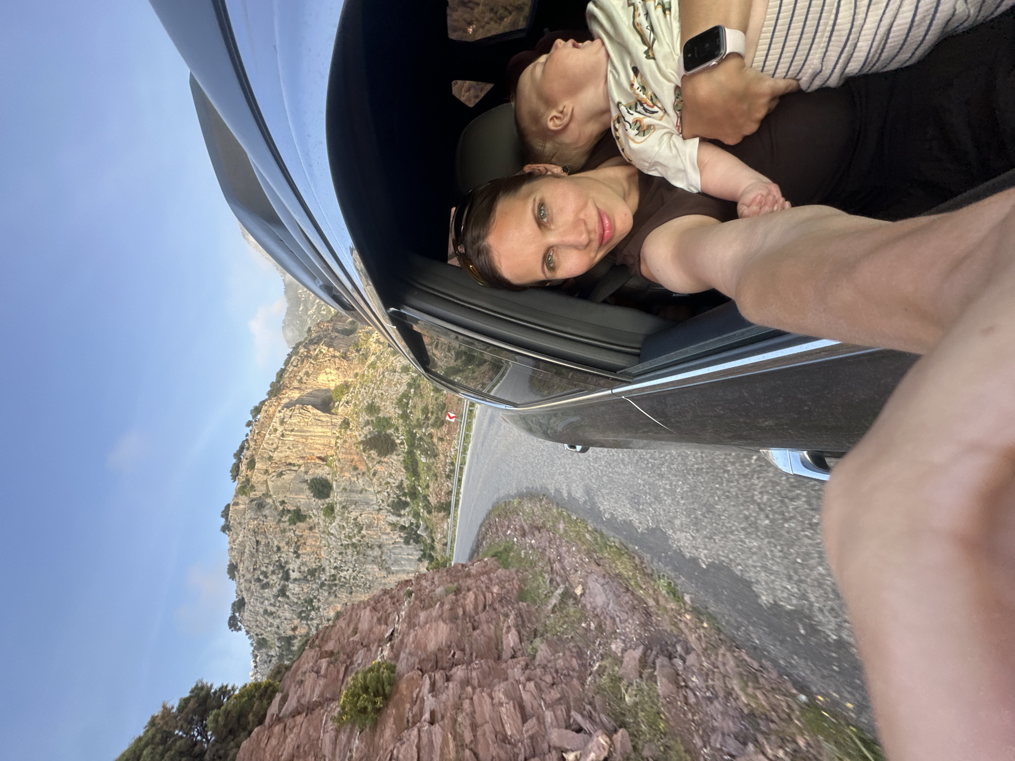 A unique selfie captures a woman and baby inside a car, looking out at a dramatic mountainous landscape in Fethiye, Turkey. The image is taken from a low angle, seemingly from the perspective of someone leaning out or holding the camera out of the car window.
In the foreground, a woman with dark hair pulled back and striking light-colored eyes smiles directly at the camera. She wears a brown top and sunglasses are pushed back on her head. Cradled in her arms is a baby or toddler, dressed in a patterned light-colored onesie, appearing calm or asleep with its head resting against her chest. A light-colored smartwatch is visible on the wrist of the arm holding the baby, and a smartphone (partially visible with a white back) is near the baby's hands. The car's dark interior, including parts of the door frame and seat, is visible around them.
Outside the car, a steep, rugged mountain dominates the background, characterized by layered rock formations and sparse green vegetation. A winding asphalt road curves alongside the cliff face, with a metal guardrail separating it from a sheer drop. Another, lower rocky formation with dense bushes is seen in the immediate foreground outside the car. The sky is clear and blue, suggesting good weather, possibly during the late afternoon or early morning when the light casts soft shadows on the mountains. A small, triangular red sign with a white center is visible on the roadside railing in the distance, but no text is discernible. The overall scene conveys a sense of travel and appreciation for the natural beauty of the mountainous region.