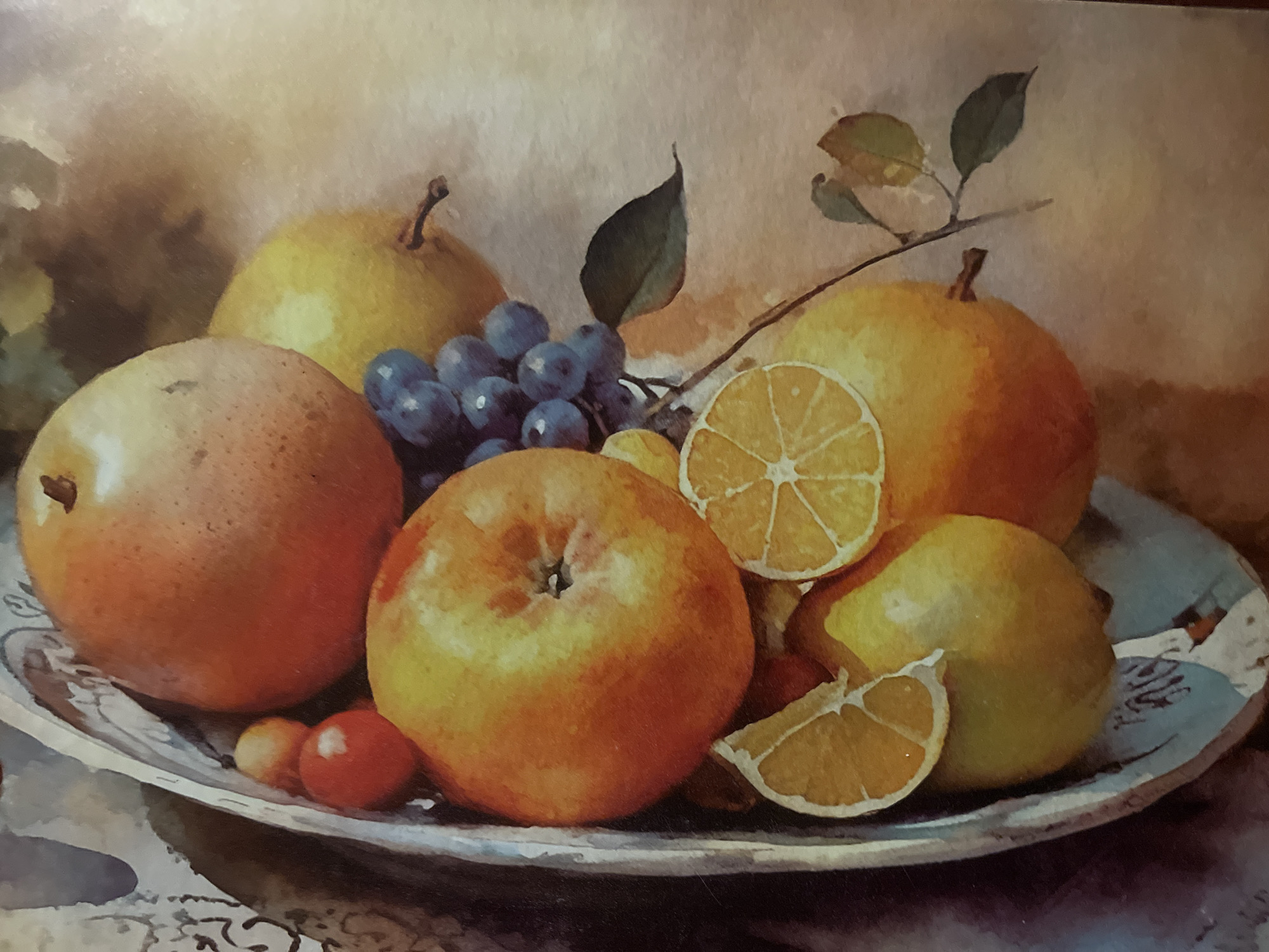 This is a still life painting of a fruit arrangement. In a decorative bowl, there are several fruits including what appear to be apples, pears, grapes, and orange slices. The fruits are rendered in warm tones of red, orange, yellow, and purple, with some green leaves from the grape stems and a separate branch with leaves. The background is a soft, muted blend of beige and brown tones, suggesting a diffused light source. There are no people, actions, or interactions depicted in this artwork. The painting does not show any location in the city of Minsk, Belarus.