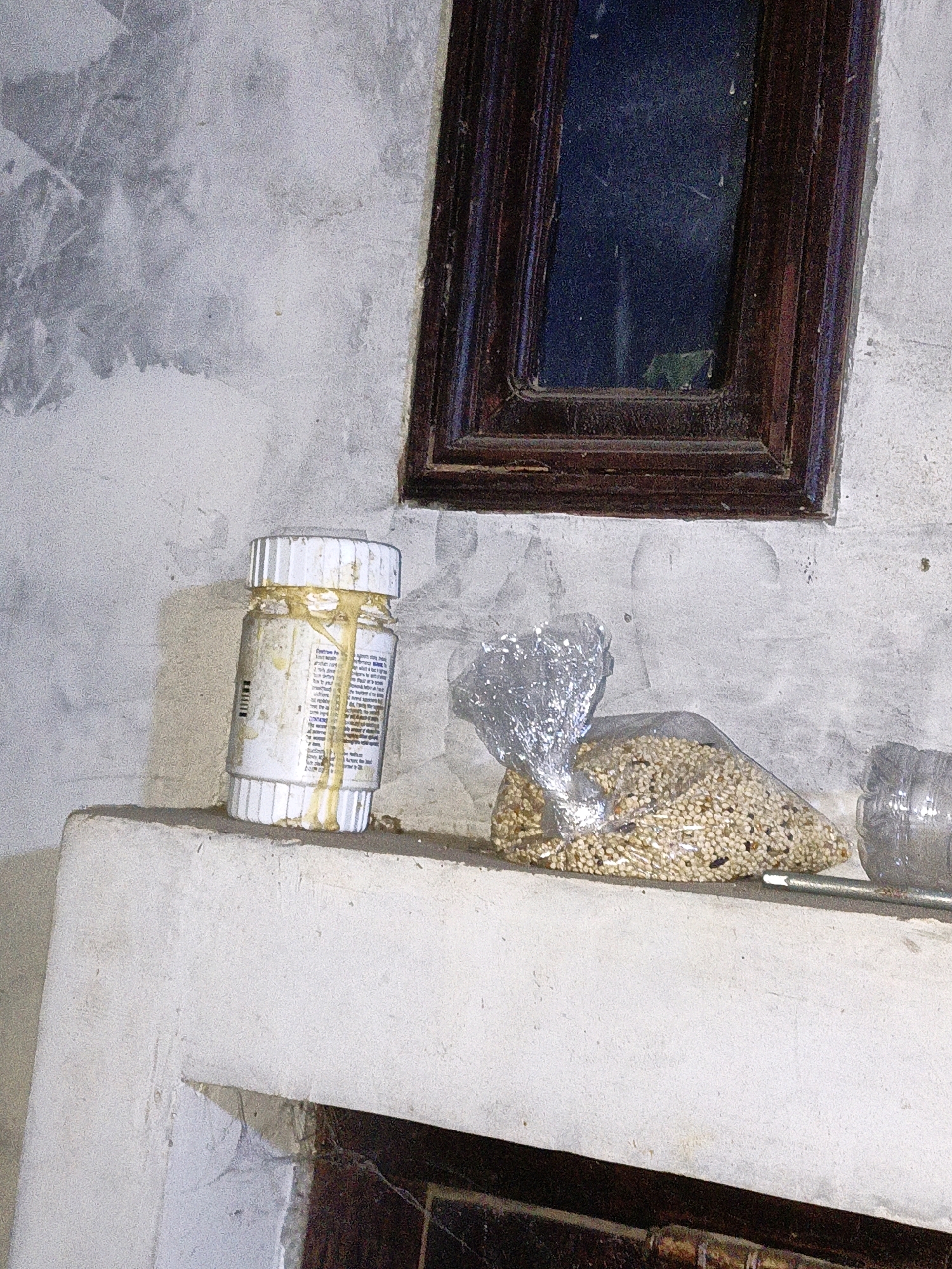 The image depicts a close-up, angled view of a weathered concrete ledge against a textured, possibly stuccoed, wall. A dark, wooden-framed window with a reflective, dark blue pane is visible in the upper right quadrant.
On the ledge, from left to right, are several objects:
1. A small, white plastic bottle with a white screw-top lid. The bottle appears to be partially filled with a yellowish substance, and there is significant yellow residue dripping down the sides of the bottle and onto the ledge. The label on the bottle is partially obscured by the residue but appears to have printed text.
2. To the right of the bottle is a clear plastic bag filled with small, light-colored seeds, some appearing to be mixed with darker particles. The bag is partially wrapped in crinkled, reflective material, possibly aluminum foil.
3. To the far right, a portion of a clear plastic bottle is visible, partially cut or damaged.
The overall environment suggests an outdoor or semi-outdoor setting, possibly a neglected area. The lighting is dim, creating shadows and highlighting the textures of the wall and objects. There are no individuals or discernible human interaction within the frame. The image does not provide any specific contextual clues to pinpoint a location within the city of Cikal, Indonesia.