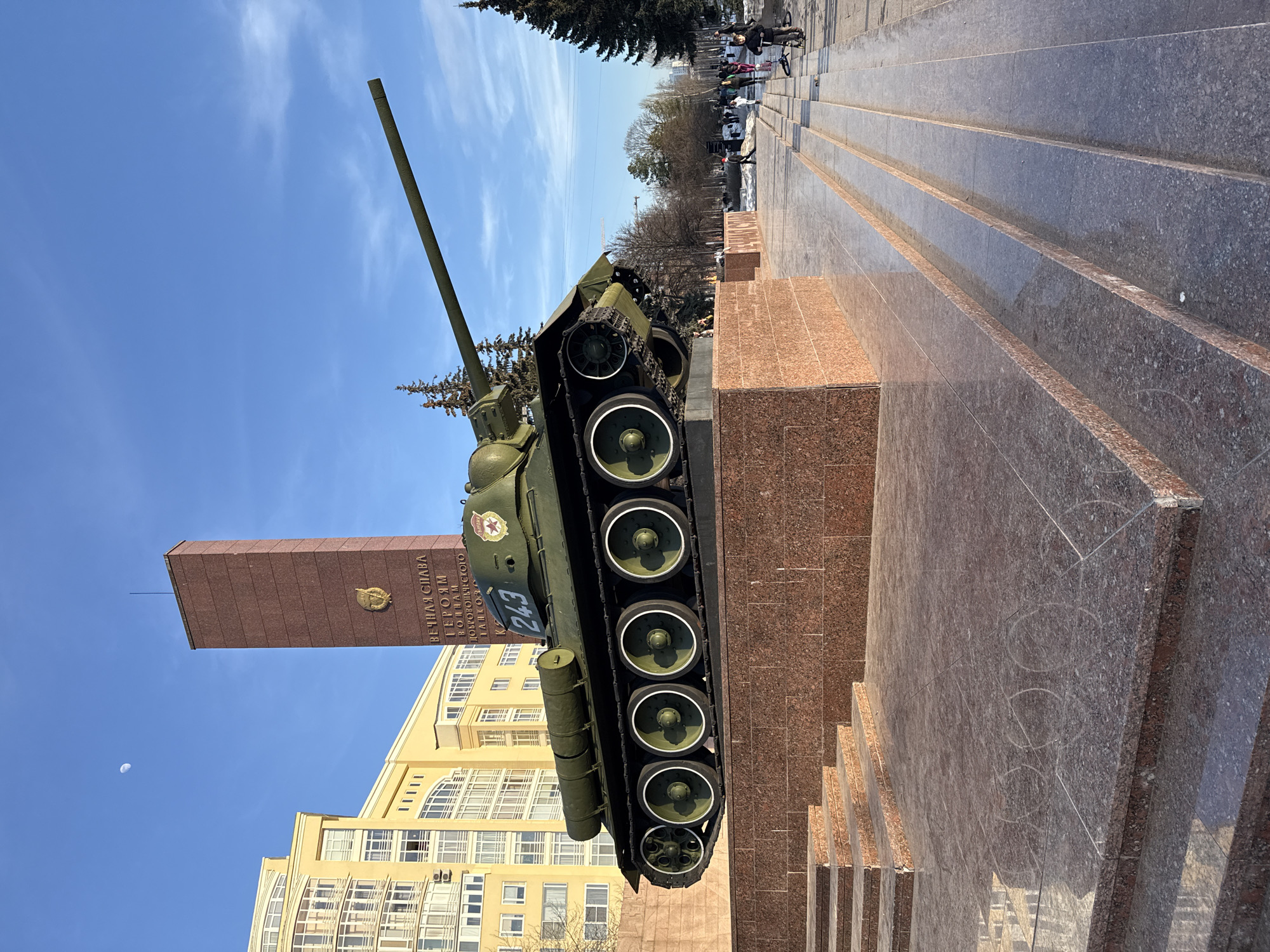 The image captures the Monument to the Ural Volunteer Tank Corps on Sibirskaya Street, Perm, Russia, under a clear blue sky with a visible crescent moon in the upper left. A green T-34 tank, bearing the number '243' in white and a red star emblem on its turret, is centrally positioned on a multi-tiered pedestal made of polished red-brown granite. Behind the tank, a tall, rectangular obelisk of matching granite stands, featuring a golden emblem and Cyrillic inscription that translates as "ETERNAL GLORY TO THE HEROES SOLDIERS OF THE VOLUNTEER TANK...". To the left of the monument, a multi-story yellow building with numerous windows is visible in the background. To the right, several evergreen trees are present, and patches of snow or ice are visible on the ground. The foreground consists of polished granite steps and a walkway, exhibiting faint circular scuff marks. Multiple individuals are visible on the walkway in the mid-ground and background; a child on a scooter is in the near right, while other pedestrians walk further away.