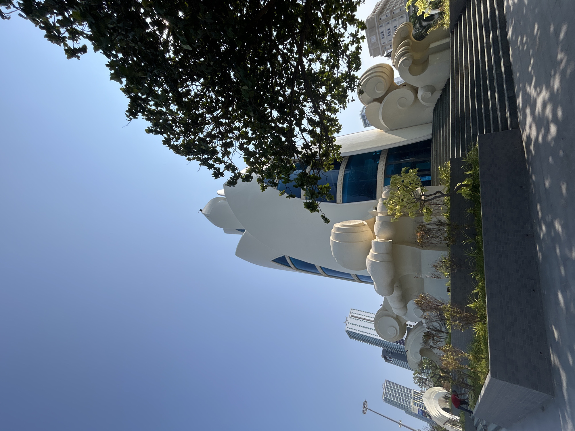 The image shows a low-angle view of the Nha Trang Center in Nha Trang, Vietnam. The building has white, sculptural elements resembling clouds or waves, with blue-tinted windows. A tree with dense foliage partially obscures the top of the building. Tall buildings are visible in the background against a clear blue sky. A pedestrian walkway or plaza with gray tile or concrete is visible to the right of the building. The photograph is taken from a perspective looking upwards and at a slight angle to the right.