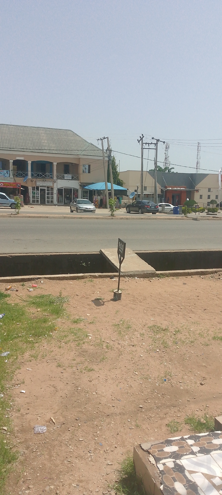 The image depicts a street scene in Jalingo, Nigeria, on a clear, sunny day. In the foreground, a patch of dry, sandy ground with sparse grass is visible, littered with some plastic bottles and debris. A concrete drain runs across the mid-ground, with a short concrete ramp extending from its edge. A signpost with "P.O.S SERVICE" printed on it stands next to the ramp.
In the background, a paved road is lined with several buildings. On the left, a two-story building features a light-colored facade with balconies and shops on the ground floor, one of which has a sign that reads "WINE STORE". Several cars are parked or driving on the road in front of the buildings. One silver car is prominently featured in the middle of the road. Further down the street, more buildings are visible, including one with a blue awning and a cluster of people gathered beneath it, possibly at a market stall or shop. Electrical poles and wires crisscross the sky, indicating the presence of infrastructure. The overall impression is of a typical commercial street on a bright, possibly warm day, with a mix of vehicles, shops, and everyday urban elements.