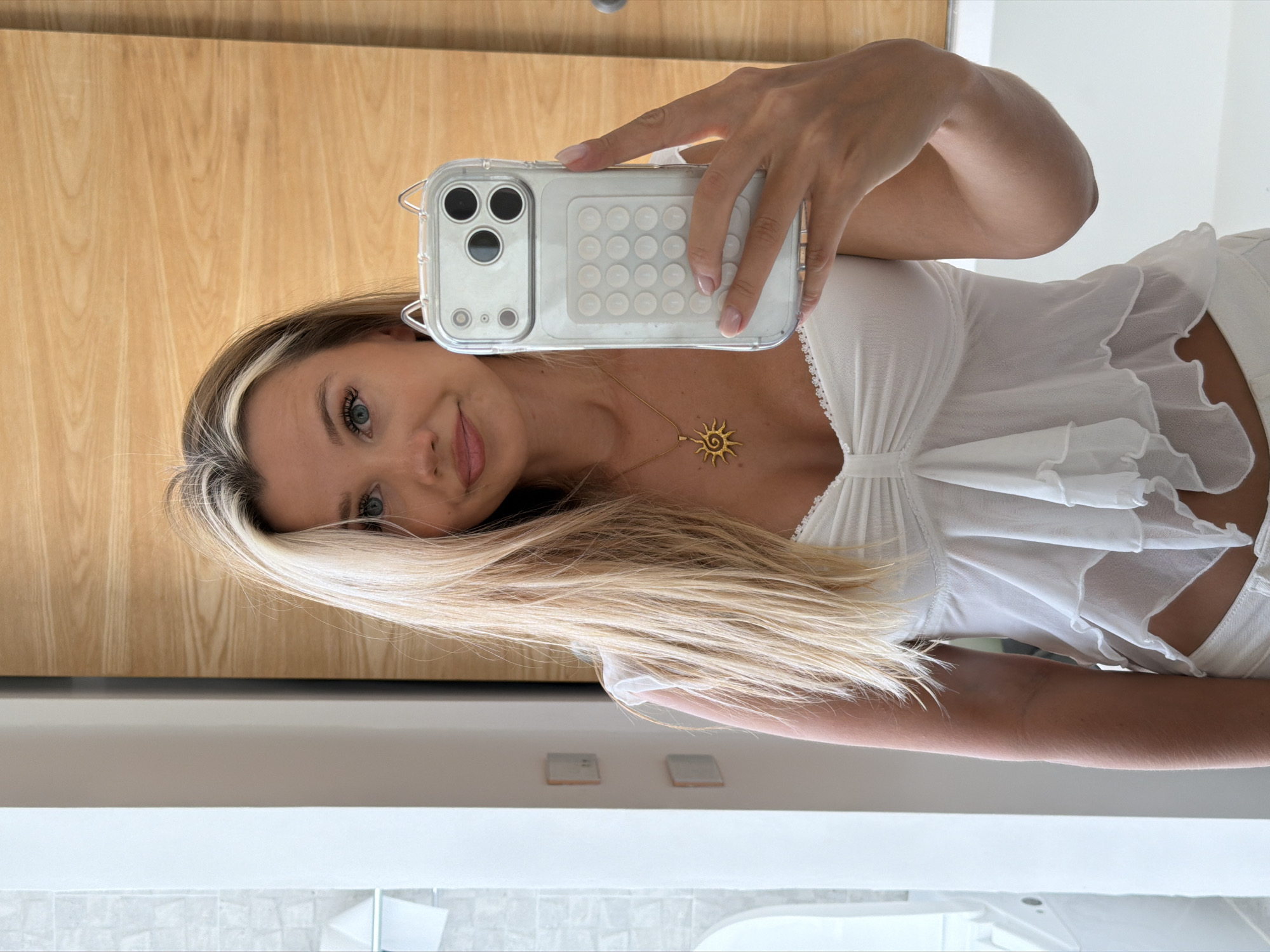 A woman is taking a selfie in a bathroom. She has blonde hair and is wearing a white top. She is holding a clear phone case with a cat-ear design. The bathroom has white tiles and a wooden door.