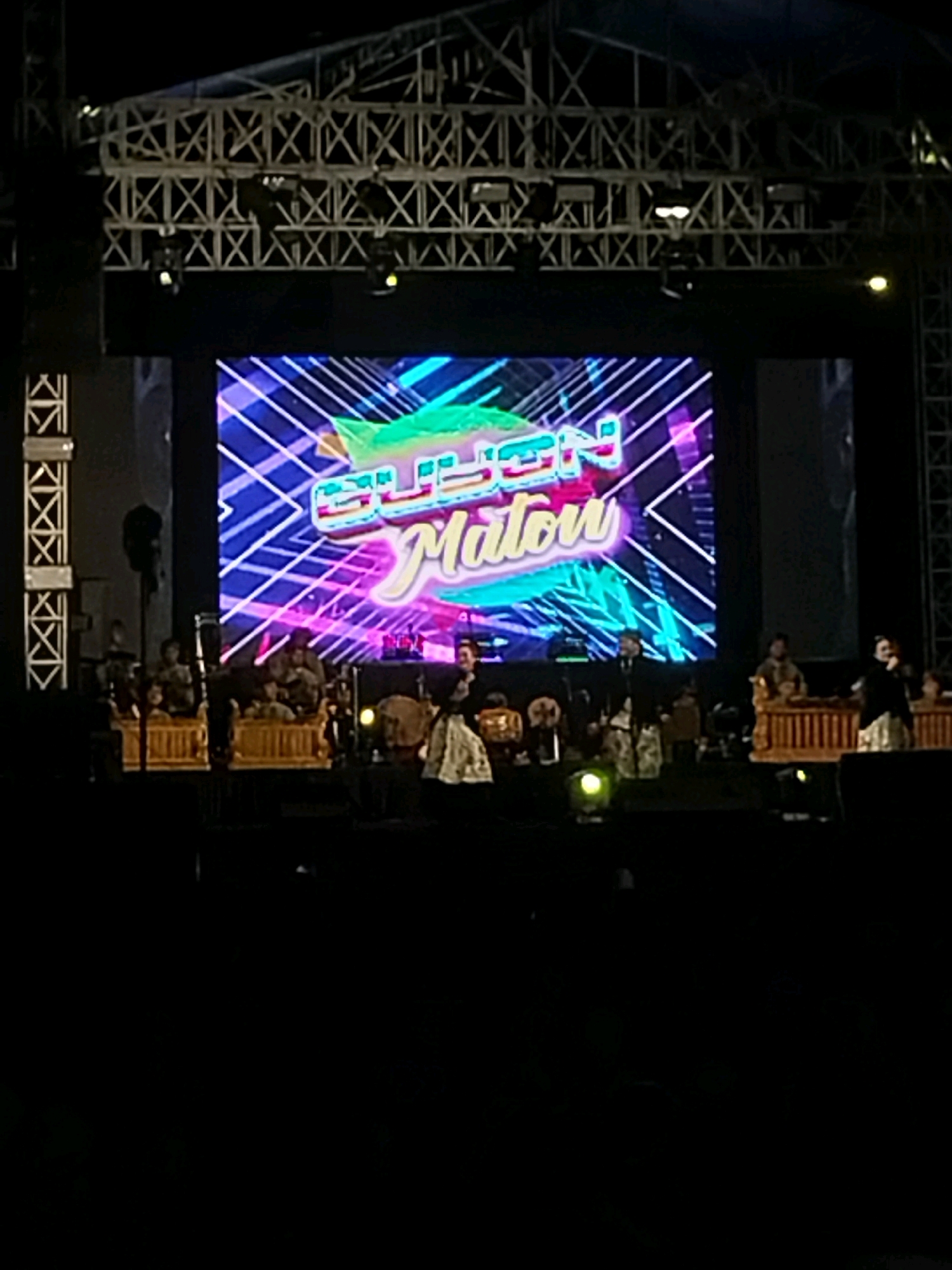 The image depicts a stage performance at night. A large LED screen displays the text "GELEN Matou" in vibrant colors against a geometric background. Below the screen, a group of musicians are seated on the stage, appearing to play traditional Indonesian instruments. Several performers are standing, some in traditional attire. The stage is illuminated by stage lights, and parts of the audience area are visible in the foreground, mostly in shadow. The overall scene suggests a cultural event or concert taking place in an outdoor or large indoor venue. Based on the information provided, the location is Nglegok, Indonesia.