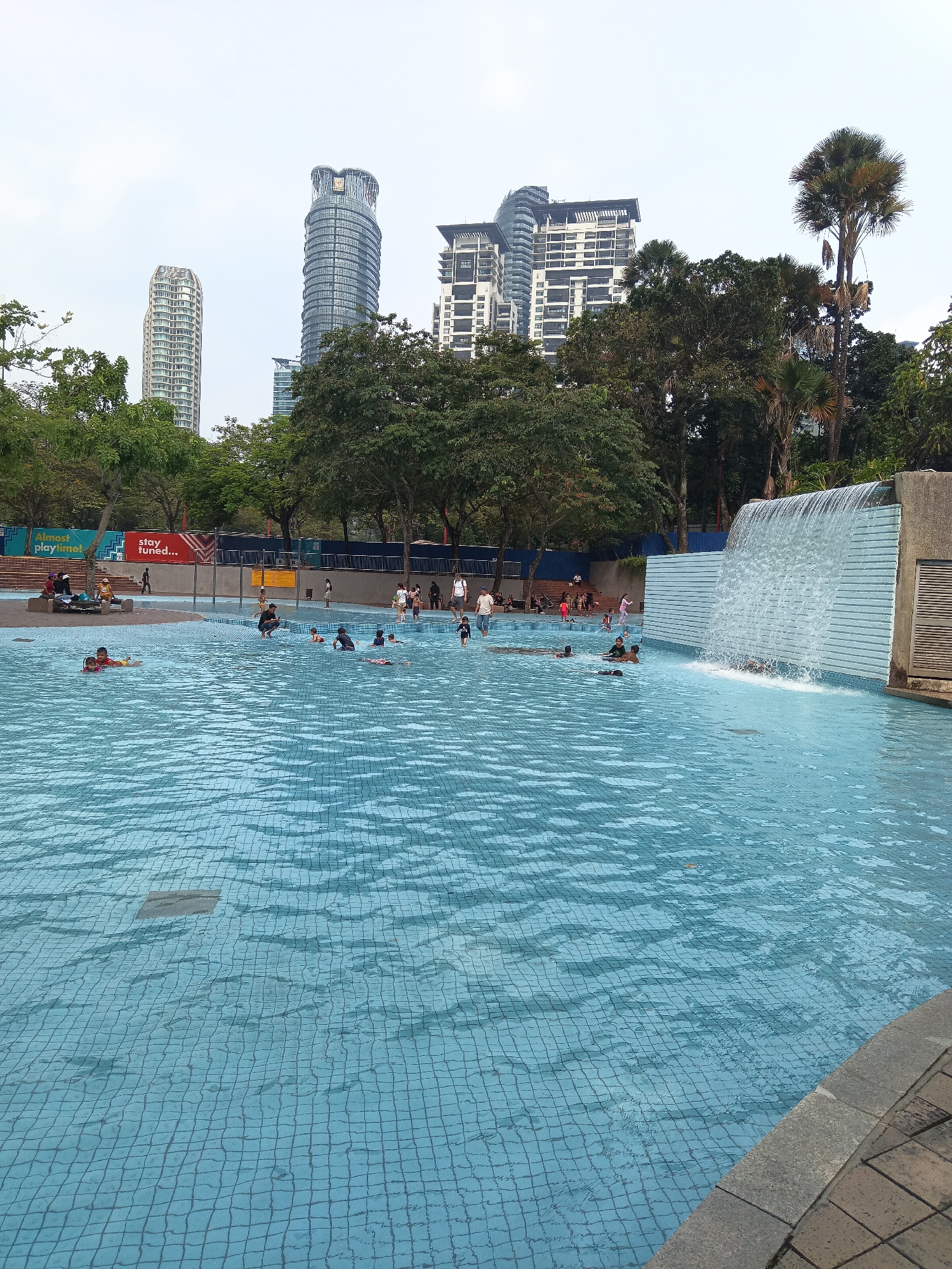 The image depicts a public swimming pool with several people enjoying the water. In the background, there is a city skyline featuring several modern skyscrapers, including the Petronas Towers, indicating the location is Kuala Lumpur, Malaysia. The pool area is tiled with blue mosaic tiles and has a waterfall feature on the right side. The environment appears to be an outdoor recreational space, likely a park, with trees and benches visible. There are adult and child swimmers in the pool, some are using flotation devices. The sky is overcast, suggesting it might be a cloudy day.
