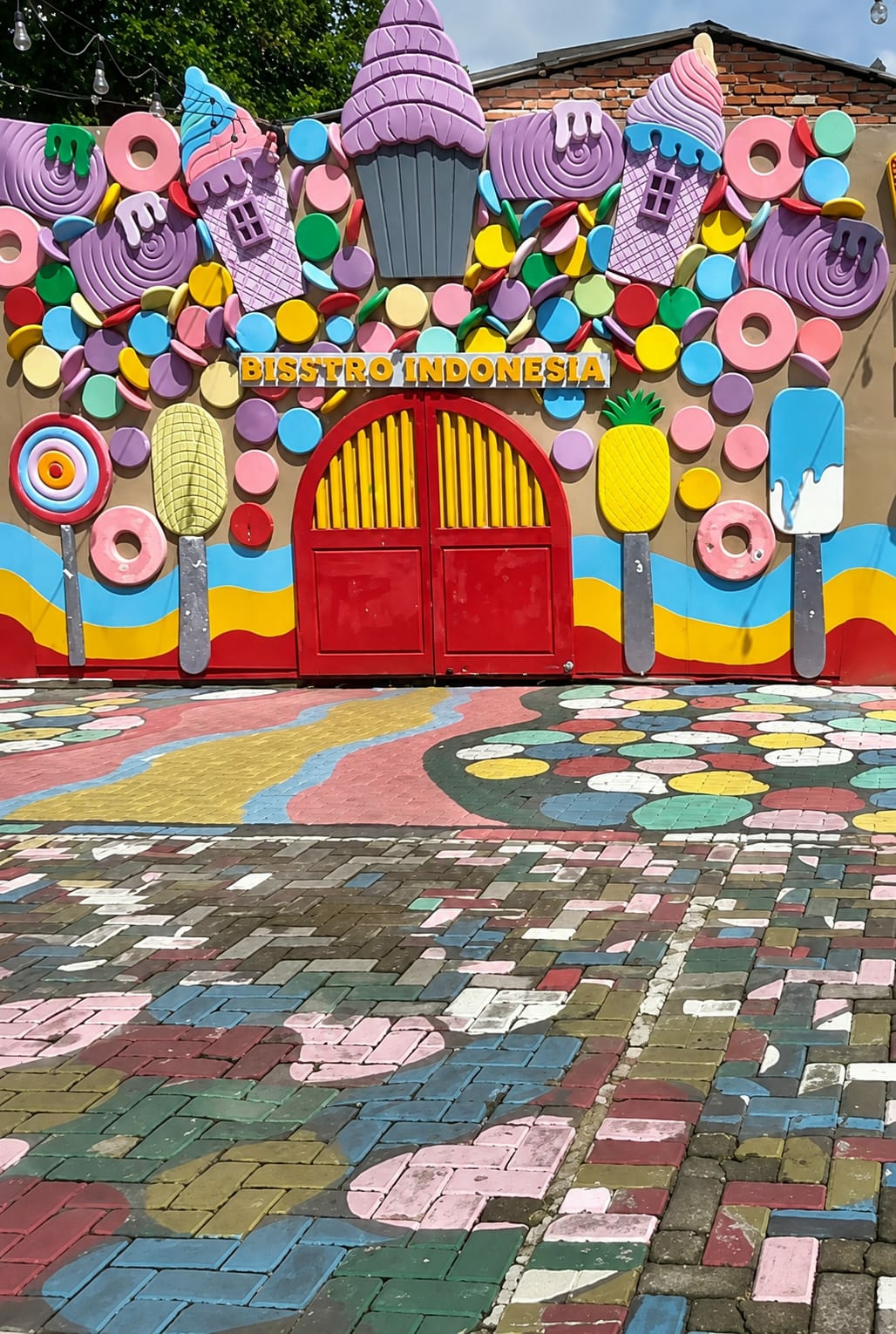 The image shows a brightly colored facade decorated with oversized candy and ice cream motifs, along with the text "BISSTRO INDONESIA" in yellow lettering. A red arched doorway with yellow vertical bars is centered on the facade. The ground in front of the facade is paved with colorful bricks painted with circular and wavy patterns. The scene appears to be an outdoor entrance to a restaurant or entertainment venue, likely located in the city of Binjai, Indonesia, given the text on the facade. The overall impression is playful and inviting, designed to attract visitors with its whimsical theme.