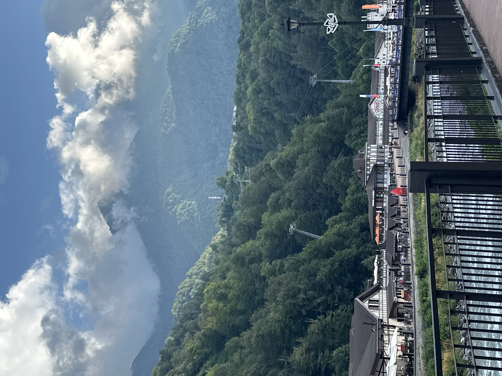 This is a vertical, eye-level, medium shot of a riverfront town nestled at the base of densely forested mountains in Estosadok, Russia. The foreground features a black metal fence and a rushing river. Behind the fence, a paved walkway runs along the river, with a row of buildings on its opposite side. These buildings have traditional Alpine-style architecture with dark roofs, white facades, and dark trim. Some have outdoor seating areas with white and orange umbrellas. People are visible walking along the walkway in front of the buildings.
Further in the background, cable car lines and towers ascend the steep, tree-covered slopes of the mountains. The mountains themselves are a dominant feature, their peaks partially obscured by dramatic, cumulus clouds against a blue sky. The overall scene conveys a sense of a popular mountain resort town, with both natural beauty and man-made infrastructure.