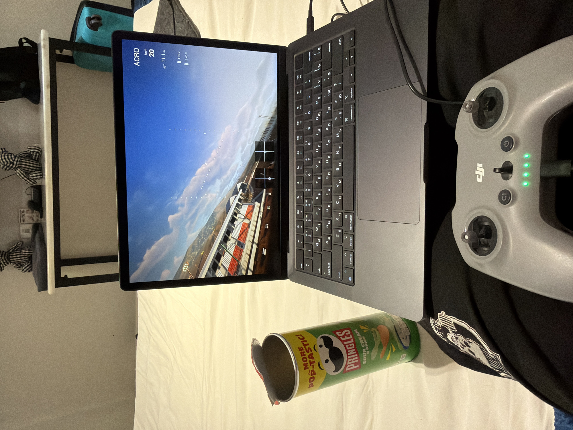 The image depicts a close-up of a person's lap with a drone controller in the foreground and a laptop open on a bed behind it. A partially opened can of Pringles chips is positioned to the left of the laptop. The laptop screen displays a first-person view of a drone flight, showing a racing track with red and white checkered barriers and a blue sky with clouds. The drone's altitude is displayed as 11.1 meters, and its speed is 20 km/h. The drone controller is grey and has various buttons and joysticks. Green LED lights indicate battery level. In the background, a white duvet covers the bed, and a shelf with decorative items and a turquoise suitcase is visible. The scene is indoors, suggesting a casual setting, possibly a bedroom or living area. The image does not provide any visual cues to indicate the location of Ban Phonsavang Neua, Laos.