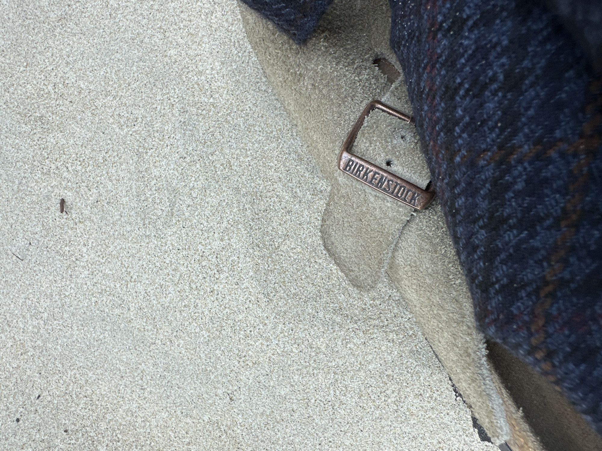 The image shows a close-up of a Birkenstock sandal lying on a surface covered in sand or fine gravel. The sandal is beige and has a copper-colored buckle with the "BIRKENSTOCK" logo engraved on it. A dark blue, possibly tweed, fabric is partially covering the sandal. A small twig is visible on the sand. There are no people depicted. The image is focused on the textures of the sand, sandal, and fabric. The location is not identifiable as Da Nang, Vietnam.