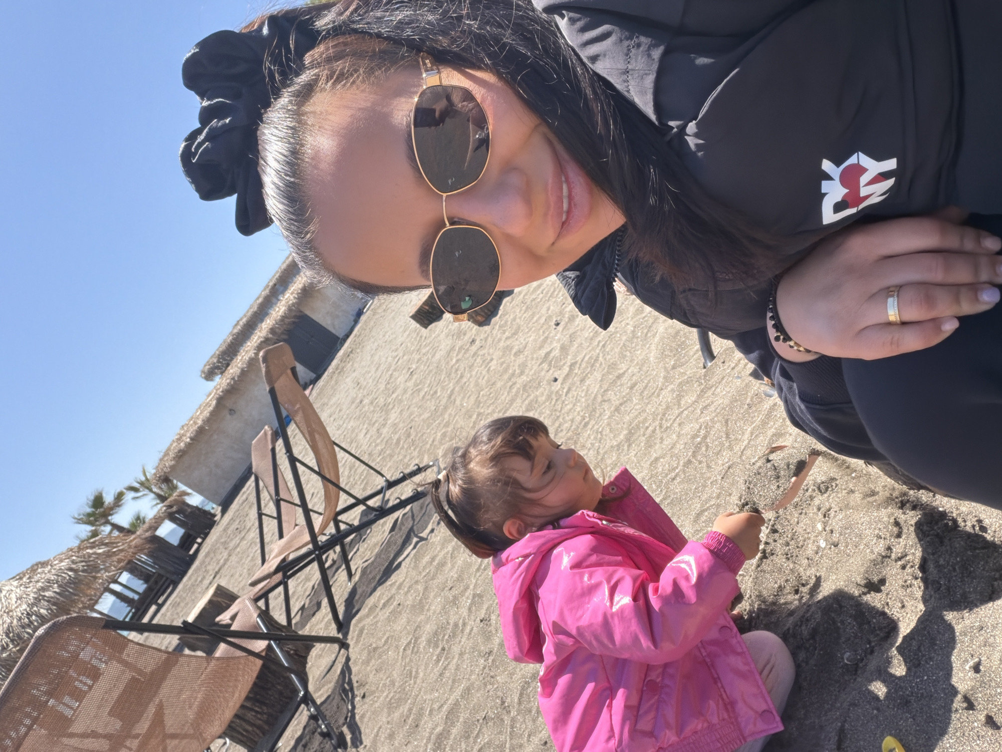 The image shows a woman and a child on a sandy beach. The woman, wearing sunglasses and a black jacket, holds the camera at an angle, capturing herself and the child in the foreground. The child, wearing a pink jacket, is sitting on the sand and appears to be playing with a leaf. Behind them, there are beach chairs and thatched-roof structures. The location appears to be a beach near a resort, somewhere in the city of Unknown, Unknown.
