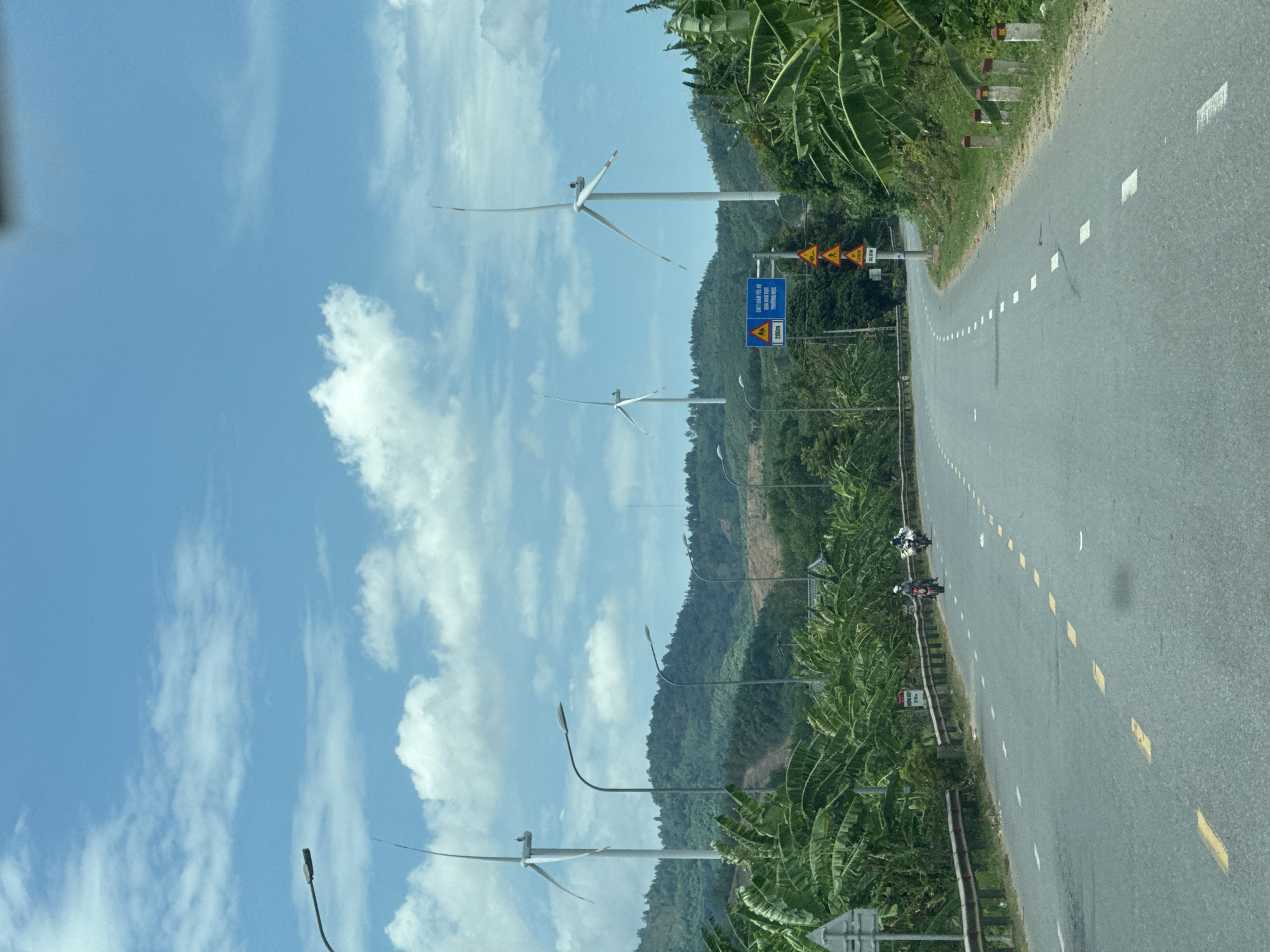 This is a landscape photograph taken from a moving vehicle on a paved road in Tân Lập Commune, Vietnam. The scene depicts a rural setting with lush green vegetation lining the right side of the road. In the background, rolling hills covered in trees rise up, and several wind turbines are visible on the slopes, indicating a wind farm. The sky is bright blue with scattered white clouds, suggesting it is daytime with pleasant weather.
The road itself is gray asphalt with white and yellow dashed lines marking the lanes. There are two motorcyclists riding on the road, heading away from the viewer. Along the side of the road, there are a few streetlights and a metal guardrail. Several road signs are visible, including yellow triangular warning signs and a blue rectangular sign with Vietnamese text. One of the visible signs reads "CHÚ Ý KHU DÂN CƯ" which translates to "Attention Residential Area" and below it "200m". Another sign appears to be a speed limit sign.
The overall impression is one of a scenic drive through a countryside area that utilizes renewable energy. The presence of the road, vehicles, and signs suggests this is an active route for transportation. The vegetation, including what appears to be banana trees, reinforces the tropical or subtropical location.