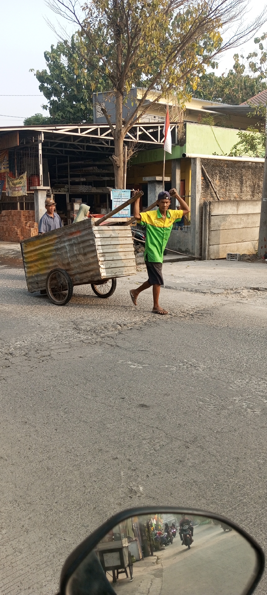 In Setu, Indonesia, on a paved road, two men are operating a large, two-wheeled cart made of corrugated metal. One man, wearing a green and yellow shirt and dark shorts, is pulling the cart by a wooden pole. He is walking with his head turned to his right, and appears to be in motion. The other man, wearing a blue collared shirt and a cap, is seated inside the cart, facing left, and smiling. The cart is laden with various items. Behind the cart, a building with a white roof and green siding is visible. In the foreground, a motorcycle's side mirror reflects a street scene with other motorcycles and small stalls. The overall environment suggests a street in a residential or commercial area.