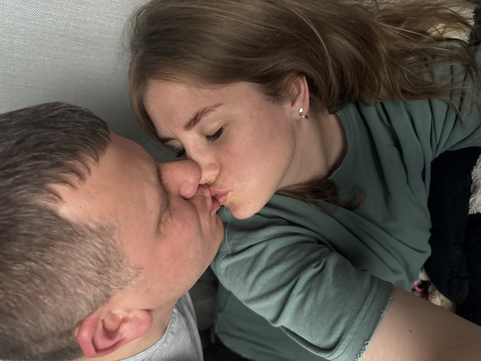 A close-up shot shows a man and a woman kissing. The man's face is turned to the left, showing his short brown hair. The woman, with medium-length brown hair and fair skin, faces the man. She is wearing a green shirt. They are lying down, and the background appears to be a textured surface. The location cannot be determined as being in Ферма, Russia, or any other specific location.