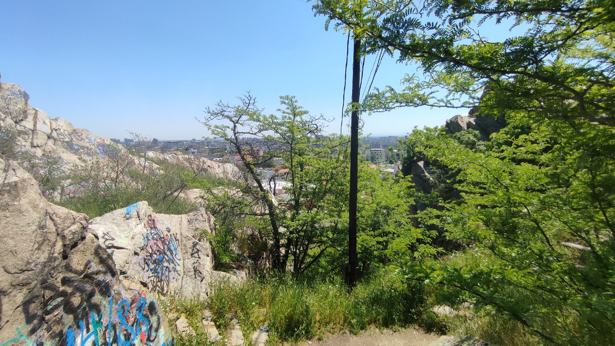 A wide outdoor shot taken on a sunny day, overlooking the city of Plovdiv, Bulgaria, from a rocky, elevated vantage point. The scene is dominated by natural elements and urban structures in the distance.
In the foreground, large, light-gray rocks covered with colorful graffiti are partially obscured by green vegetation. A few small trees with fresh green leaves stand amidst the rocks. A utility pole with visible wires is located in the middle ground.
Beyond the rocks and trees, the cityscape of Plovdiv spreads out under a clear blue sky. Buildings of various heights and architectural styles are visible, giving a sense of the city's density.
The overall impression is one of a natural, slightly wild area overlooking an urban environment, suggesting a location on the outskirts of the city or within a natural park on a hill.