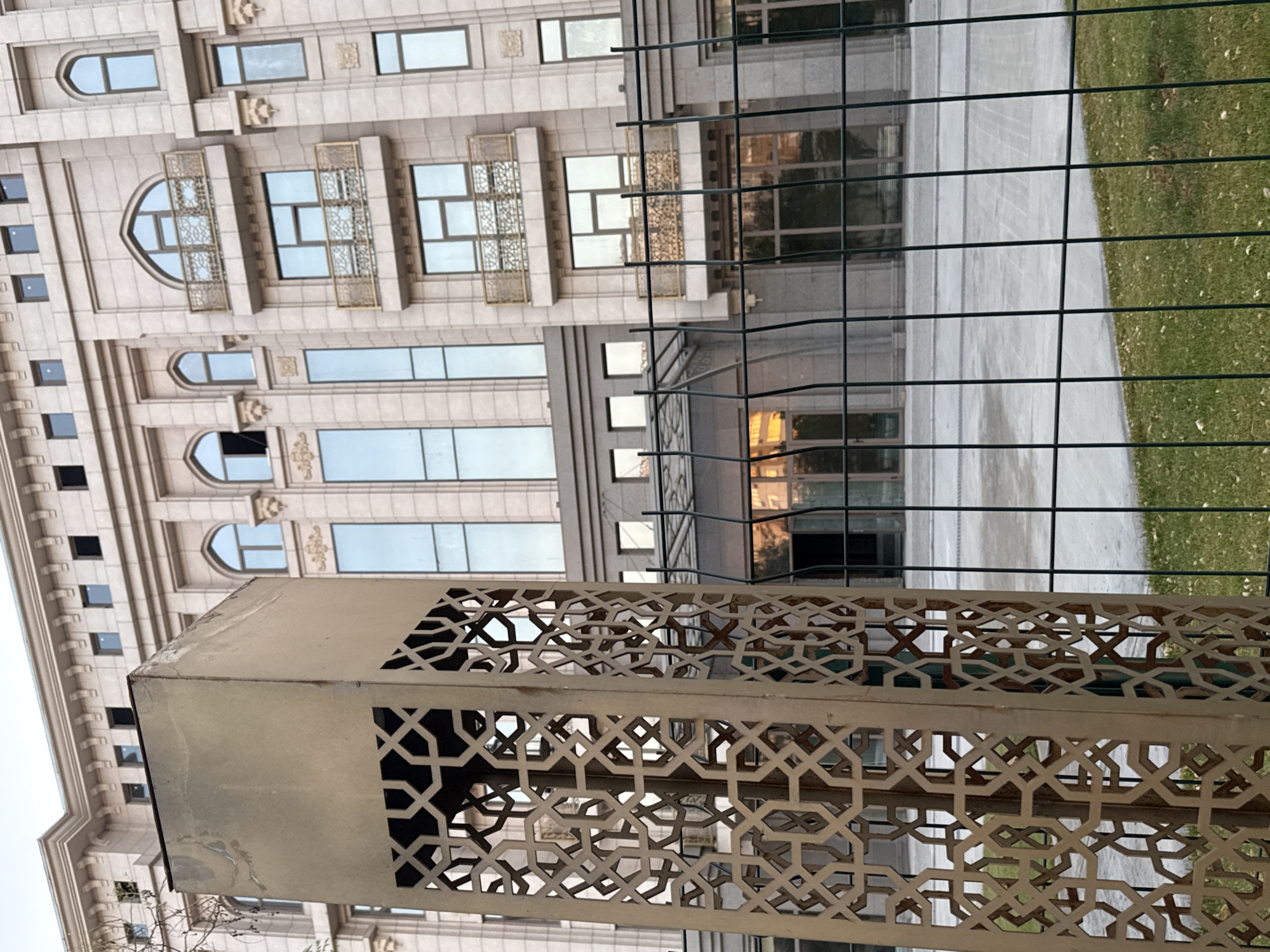 An outdoor, eye-level, medium shot shows the facade of a light beige, multi-story building with arched windows and decorative balconies. In the foreground, a metal structure with an intricate geometric cutout pattern stands in front of a green wire fence. The building's architecture suggests a modern interpretation of traditional Islamic design, possibly a residential or commercial complex. The scene is set in an urban environment, with the building occupying most of the background. The time of day appears to be late afternoon or early evening, as indicated by the warm light reflecting on the building's windows. No people are visible in the frame. This building is located on A. Kodiriy Street in Tashkent, Uzbekistan.