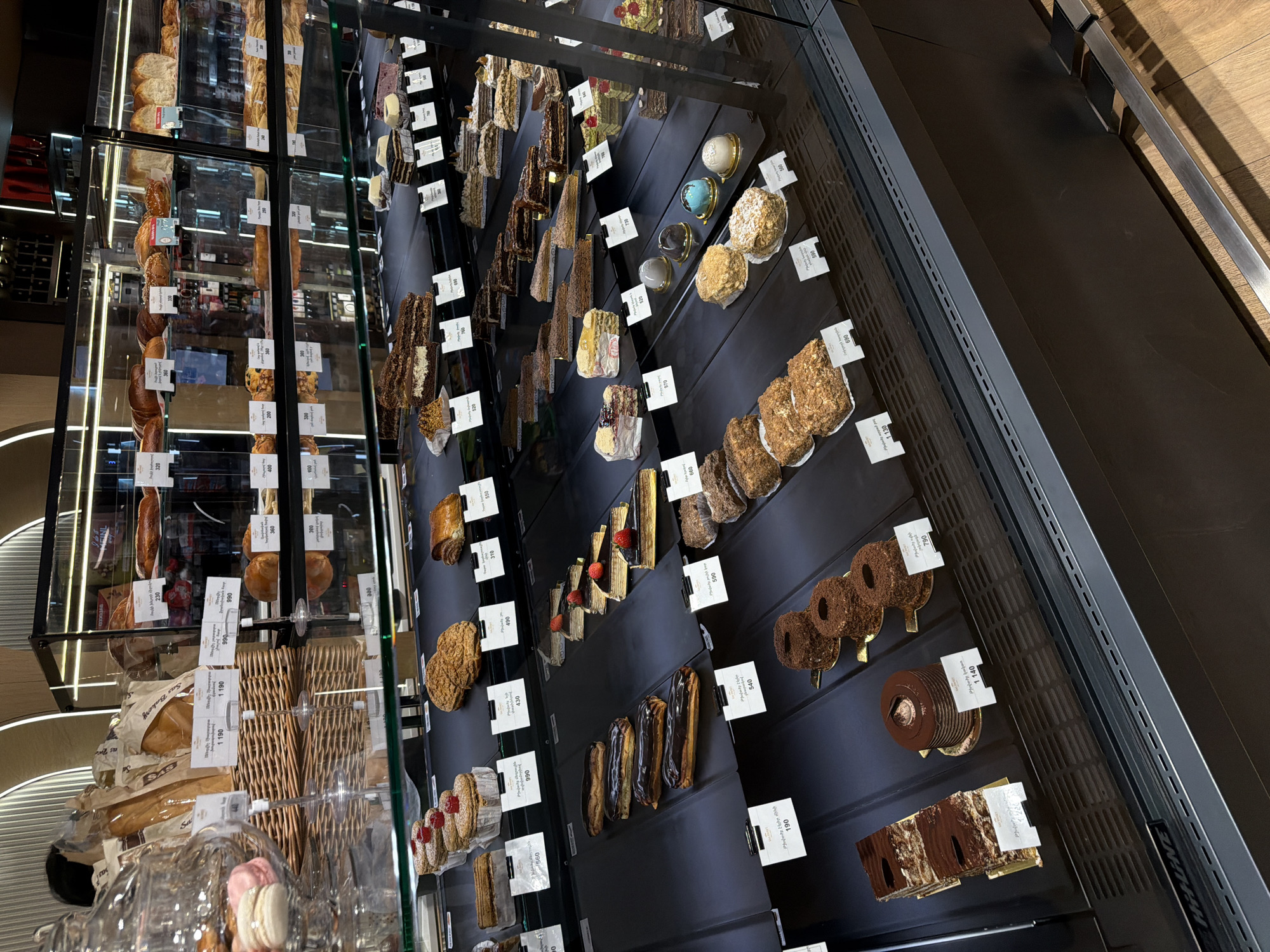 The image depicts a display case filled with various baked goods and pastries. The case is likely located in Yerevan, Armenia, judging by the language on the price tags. The left side of the case showcases a variety of bread and rolls arranged on shelves. The right side displays an assortment of cakes, tarts, and eclairs, each with a small price tag. The arrangement is orderly, with items neatly aligned on black shelves. The environment appears to be a bakery or pastry shop.