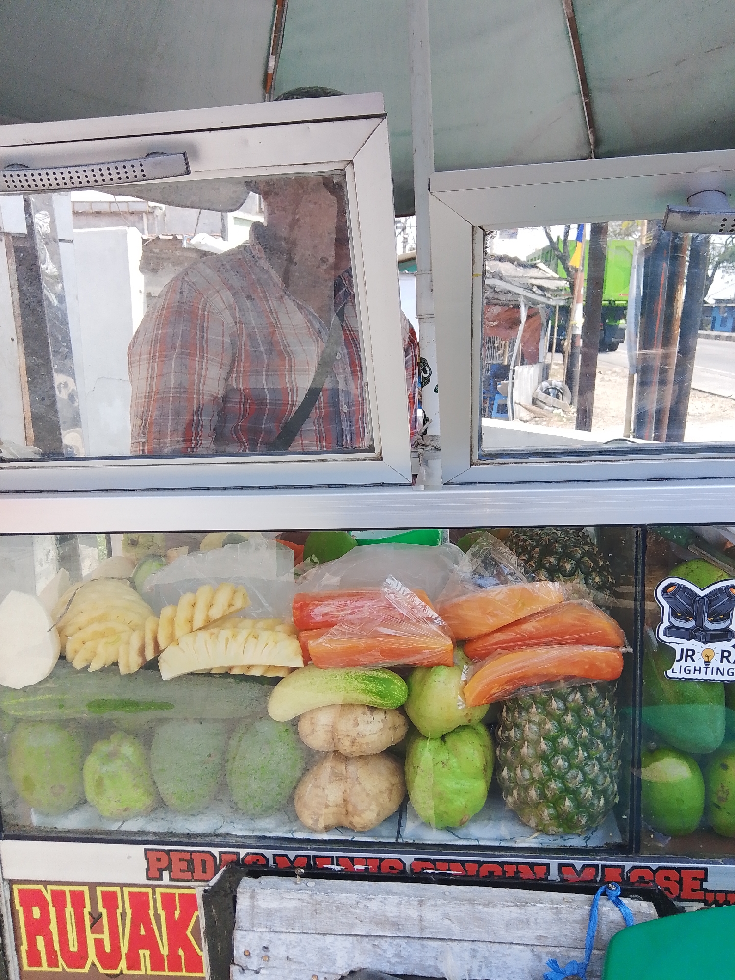 The image shows a fruit stand with various fruits displayed in a glass case. The fruits include sliced pineapple, whole pineapples, papayas, guavas, cucumbers, and what appear to be jicama root slices. Some fruits are individually wrapped in plastic. A person wearing a plaid shirt is reflected in the glass case, suggesting they are the vendor or a customer. The stand has a sign with the word "RUJAK" in red and yellow letters, indicating it sells rujak, a traditional Indonesian fruit salad. The stand appears to be located outdoors, with a road and buildings visible in the background, consistent with a street-side vendor in Sedo Lor, Indonesia. The overall scene depicts a typical street food vendor setup.