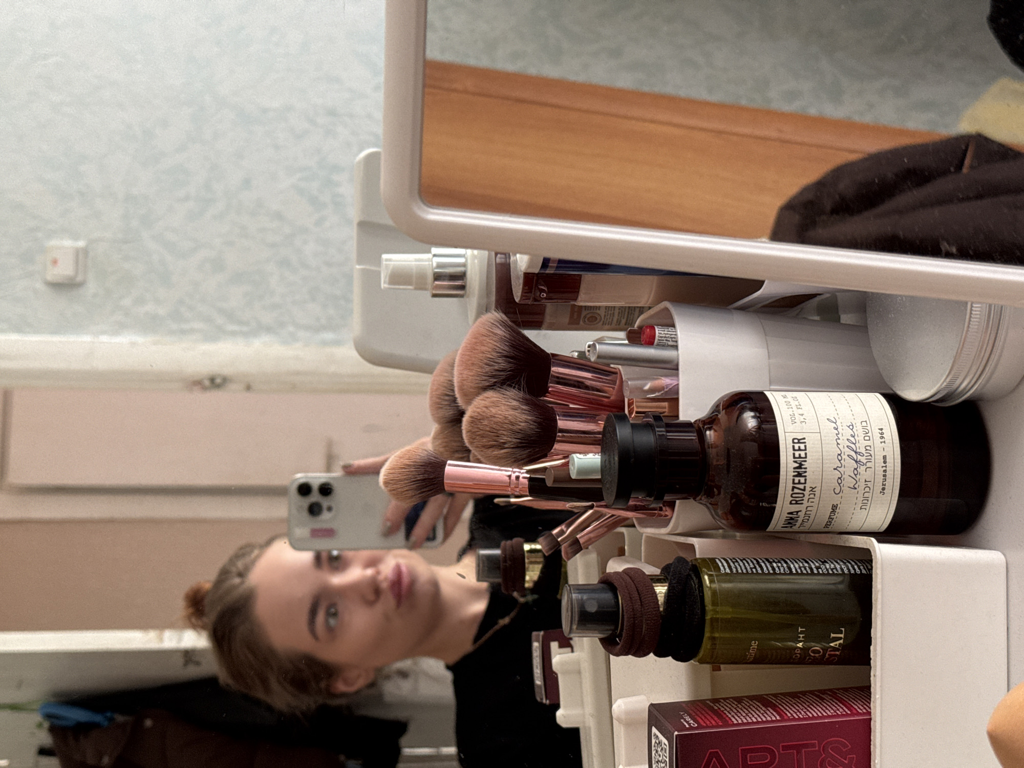The image depicts a reflection of a person holding a smartphone, likely taking a selfie, with a storage organizer unit containing various cosmetic items in the foreground.
The items within the organizer include:
* Makeup brushes
* A dark brown glass bottle labeled "ANNA ROZENMEER" with the words "caramel waffles" and "Jerusalem - 1964", likely a perfume.
* A green glass bottle with partially visible label "РО... STAL"
* A box labeled "APTS & CARELY".
The organizer is white and seems to be tiered, with multiple shelves. The background includes a wall with a textured pattern and an electrical outlet.
The location in the city of Chaikovsky, Russia cannot be discerned from the image.