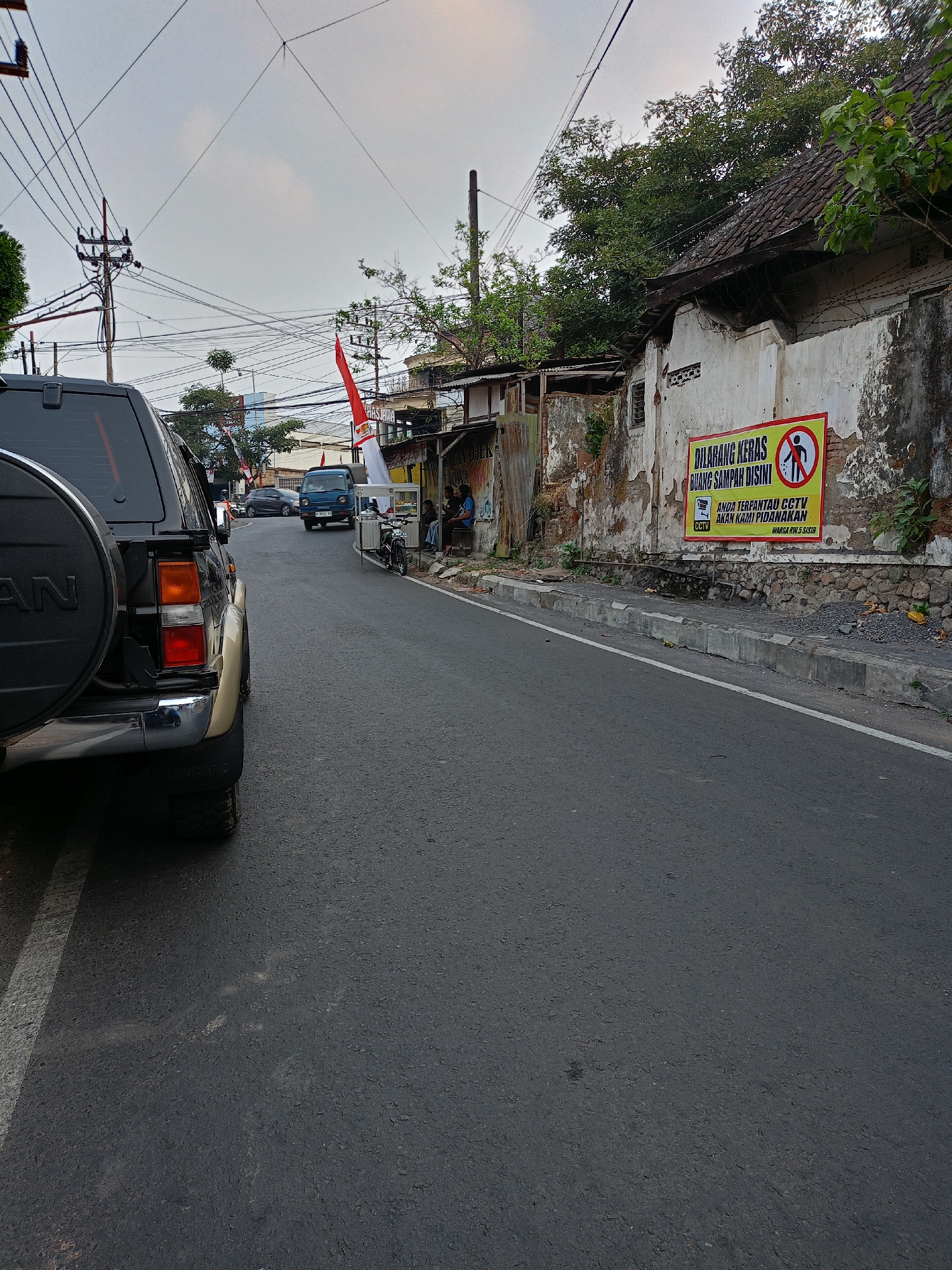 The image shows a street scene in Batu, Indonesia. In the foreground on the left is the rear portion of a dark-colored SUV, its spare tire mounted on the back. The vehicle is parked alongside a road with a white line marking a parking space. The road itself is asphalt.
On the right side of the road, a weathered white wall is visible, with signs of peeling paint and some damage. A large, yellow sign with red text is prominently displayed on this wall. The sign reads "DILARANG KERAS BUANG SAMPAH DI SINI" (Strictly Forbidden to Throw Trash Here) and includes a circular icon with a prohibited trash symbol. Below this, it states "ANDA TERPANTAU CCTV AKAN KAMI PIDANAKAN" (You are monitored by CCTV, we will prosecute) and "WARGA RW.5 SISIR".
Further down the street, there are several people sitting on a bench in front of a small food stall or cart. A blue truck is driving away from the viewer in the middle of the road. Indonesian flags, red and white, are visible on poles in the distance, suggesting a national holiday or event. Trees and power lines are present throughout the scene, and the sky is overcast. The overall impression is of a residential or commercial street in a somewhat aged urban environment.