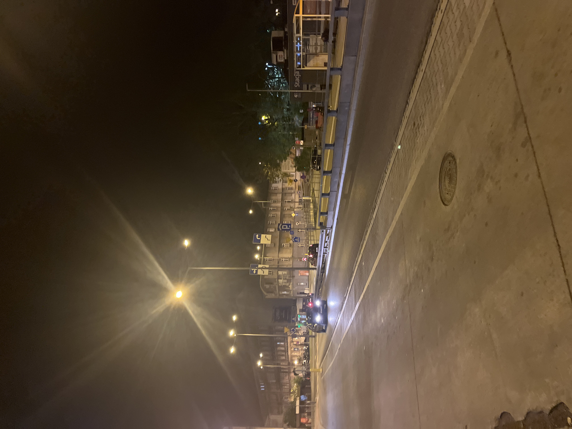The image captures a nighttime street scene in an urban area. Streetlights illuminate a road, lined on one side by buildings, trees, and signage. Cars are present on the road, with their headlights visible. There is a sidewalk or paved area beside the road. The sky is dark, indicating it is night. A structure with the word "Stacja" (station) visible is present in the distance.