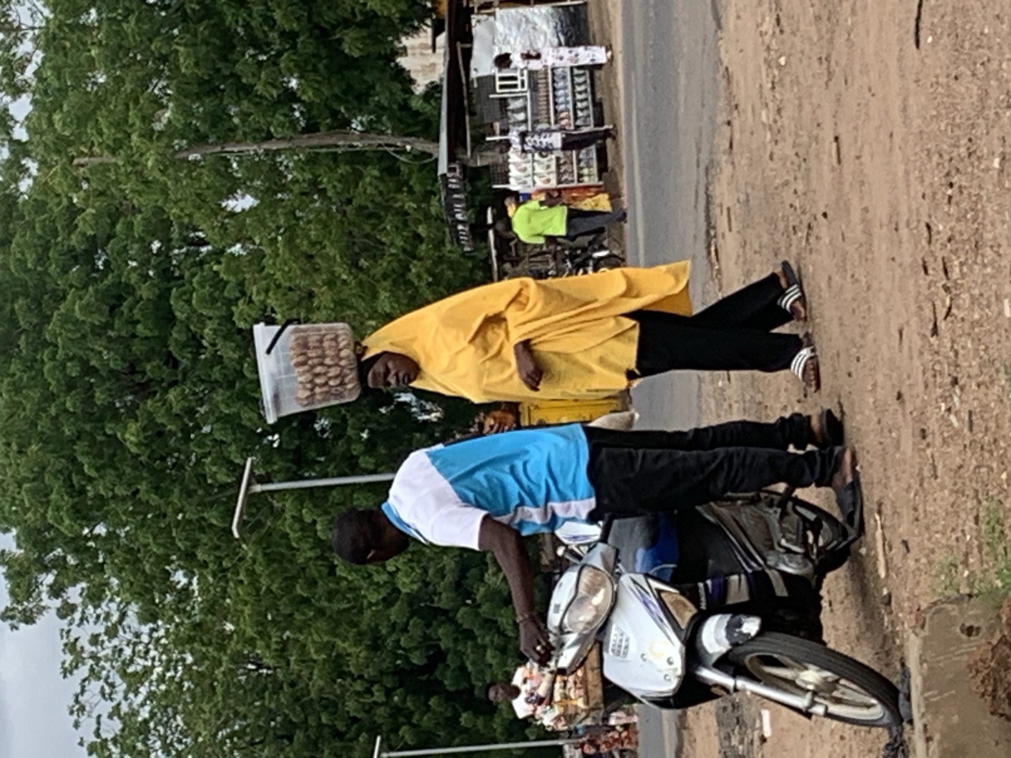 This image captures a daytime street scene in Shendam, Nigeria. In the immediate foreground, a dark-skinned man wearing a white and blue short-sleeved polo shirt, black pants, and brown sandals stands to the left of a silver and blue scooter-style motorcycle. He holds the handlebars of the motorcycle, which is parked on a reddish-brown dirt and gravel shoulder. To his right, a dark-skinned woman, dressed in a bright yellow head covering and matching flowing outer garment over black pants, walks towards the right. She balances a clear plastic bucket filled with numerous light-brown, oval-shaped items (likely nuts or seeds) on her head. Her feet are clad in striped slide sandals.
In the midground, a paved road separates the foreground figures from a row of makeshift market stalls on the opposite side. These stalls are constructed from corrugated metal and wood, displaying various products. Several individuals are visible near these stalls, including a person in a bright green shirt, another in a patterned shirt, and a third in a white and patterned dress. Further left in the midground, another dark-skinned man in a white shirt is partially visible, attending to a wooden cart filled with items. Two modern streetlights, each with a flat, square solar panel, stand on tall poles on the left and center-right portions of the frame. The background consists of dense green foliage from numerous trees, indicating a lush environment under an overcast sky.