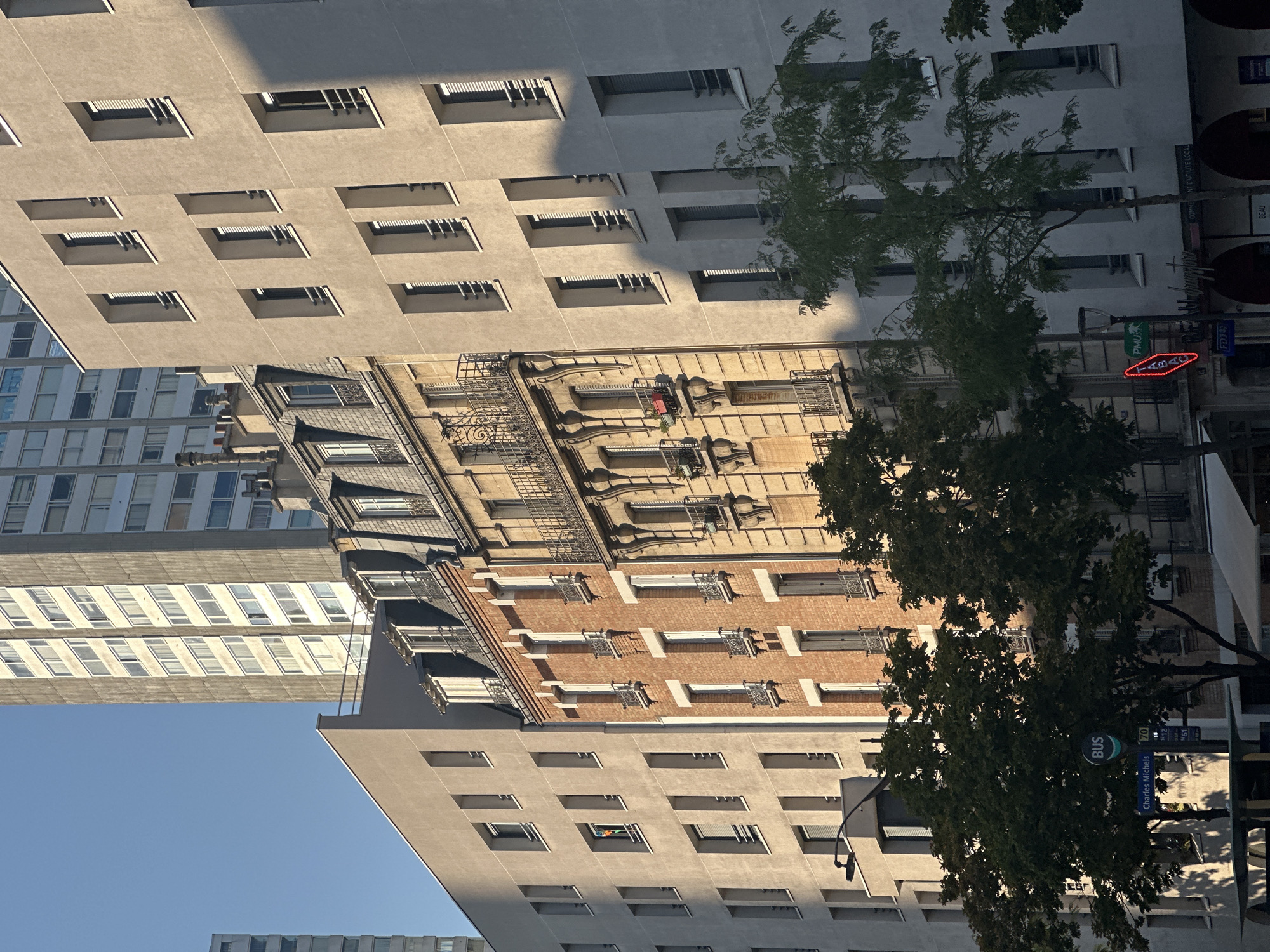 The image captures a street scene in Paris, France, focusing on a row of multi-story buildings. The architecture varies, with a modern, light-colored apartment building on the left, a classic brick building with ornate balconies and mansard roofs in the center, and another modern, light-colored building on the right. Sunlight casts distinct shadows across the facades, indicating it is likely late afternoon.
In the foreground, trees with dark green foliage partially obscure the lower levels of the buildings. A blue bus stop sign with white lettering is visible on the left, along with a street sign reading "Charles Michels." A red neon sign advertising "TABAC" is present on the right side of the street. The sky is clear and blue. There are no visible people or distinct actions taking place. The overall impression is of an urban environment with a mix of architectural styles and the characteristic shadows of a sunny day in a European city.