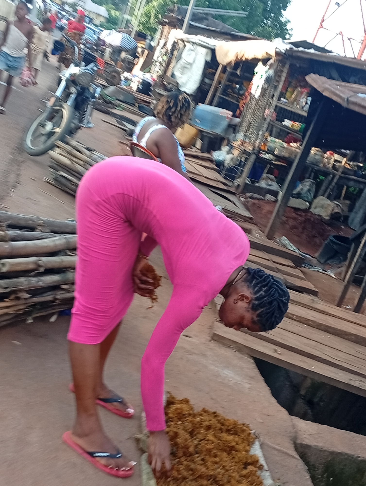 A woman in a vibrant pink bodycon dress is bent over, sorting through a pile of brown, dry material on the ground in Nsugbe, Nigeria. She has dark skin and her hair is styled in black braids. Her red flip-flops are visible on her feet. To her left, a stack of bamboo-like poles leans against a dirt surface. In the background, a motorcycle is parked, and other people are visible amidst market stalls constructed of wood and corrugated metal. One person in the background wears a white top with blue patterns. The scene appears to be an outdoor market setting.