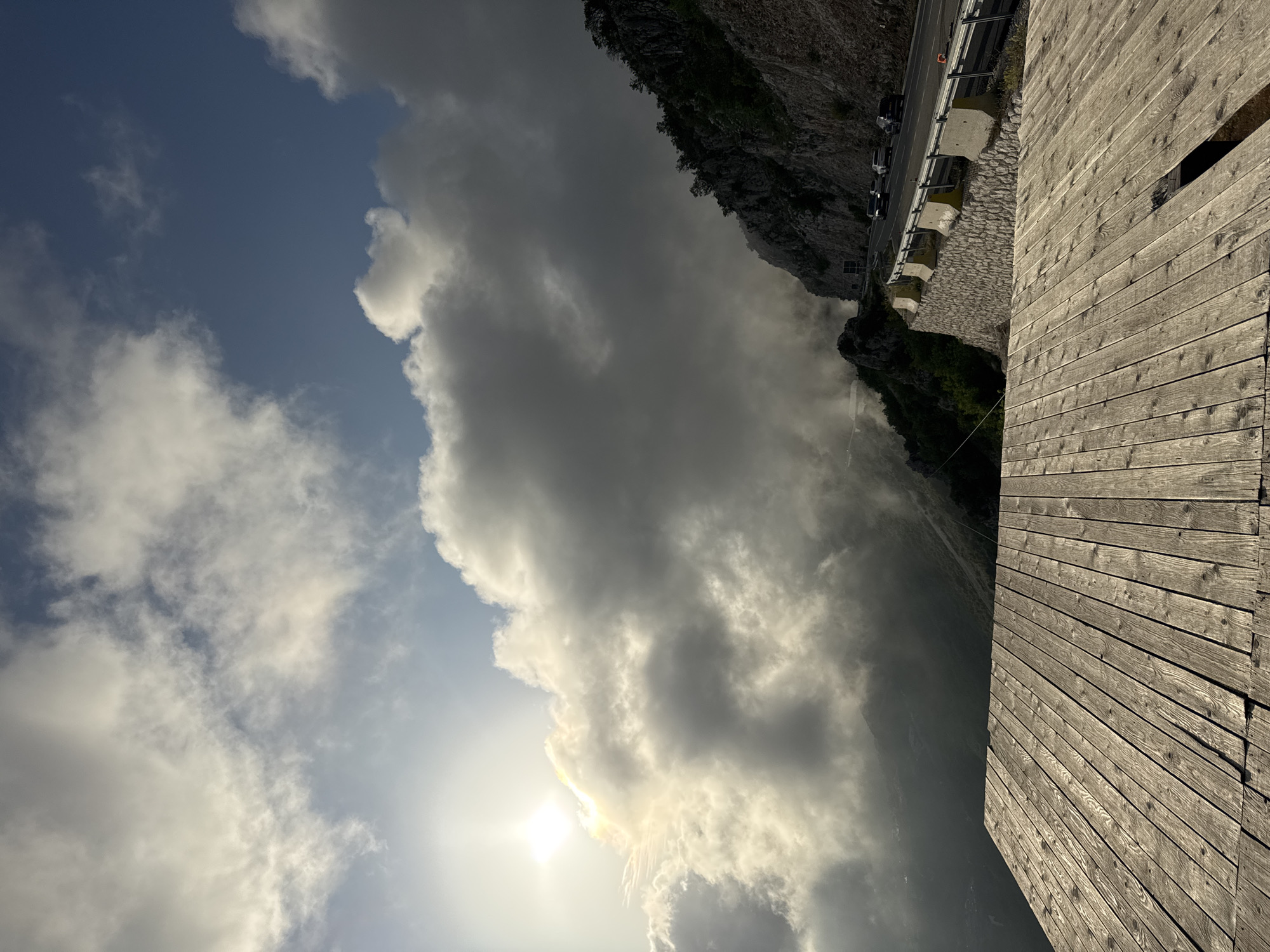 The scene captures a view in Brajici, Montenegro, featuring a combination of natural and man-made elements. The sky dominates the upper left portion of the frame, filled with a mix of bright blue patches and thick, gray clouds, with the sun shining faintly behind them. A road with several parked cars runs alongside a stone wall and vegetation along a rocky cliff face. The right side of the image features a weathered wooden structure with visible planks. The perspective suggests the photo was taken from a slightly elevated position, perhaps from a platform or viewing point.