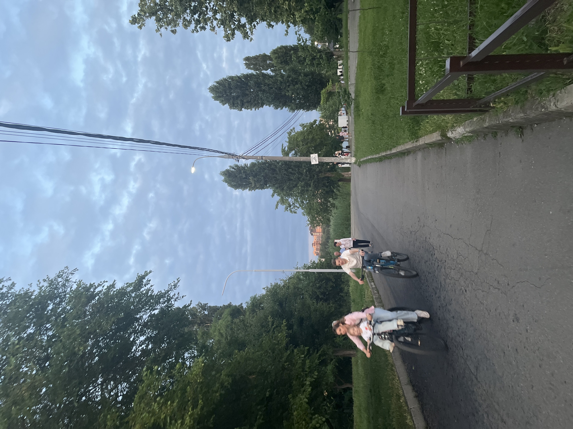 This media file shows a group of people cycling on a paved path in a park-like setting. The sky is overcast with scattered clouds, suggesting it might be late afternoon or early evening. Lush green trees line the path on both sides, and a grassy area with some wildflowers is visible to the right, bordered by a low concrete curb and a dark brown metal railing. In the distance, several trees form a backdrop, and hints of buildings can be seen beyond them.
The main subjects are several individuals on bicycles. In the foreground, two young people are riding a tandem bicycle. The person in the back is wearing a pink hoodie and jeans, while the person in the front appears to be wearing a white t-shirt and jeans. Further up the path, another person is riding a bicycle, wearing a light-colored t-shirt and jeans. Two more figures are visible further in the distance, also appearing to be on bicycles. The overall mood seems casual and relaxed, suggesting a leisurely activity on a pleasant day. The lighting indicates it is not midday. There are no visible text elements in the image. The location context suggests this scene is in Krasnodar, Russia.