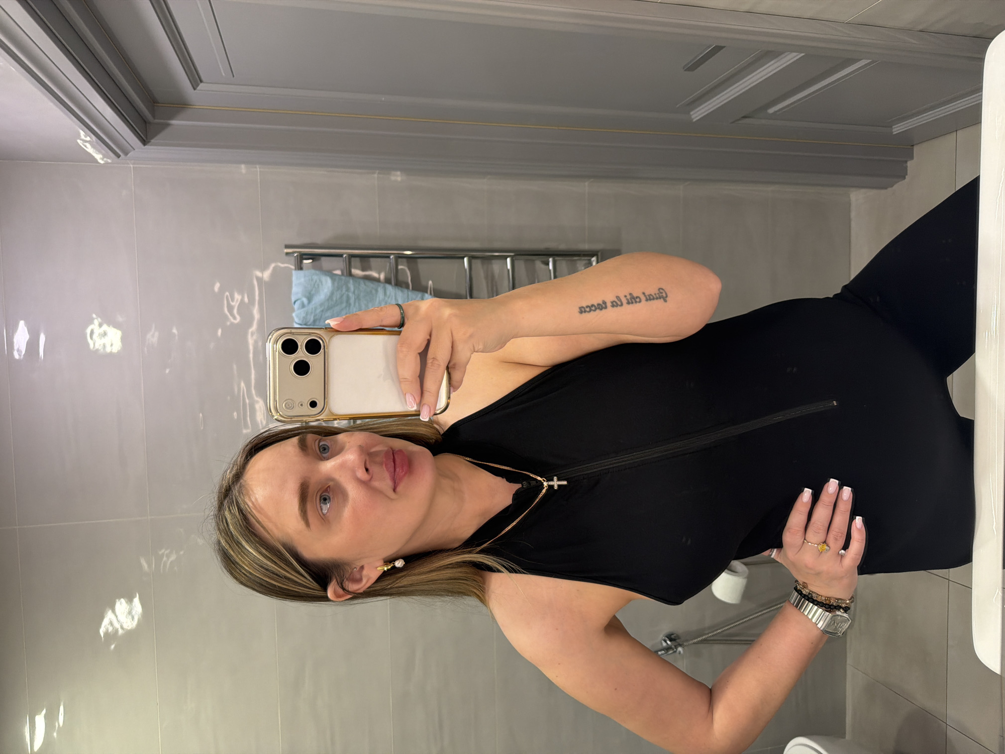 A woman is taking a selfie in a bathroom. The woman has blonde hair and is wearing a black sleeveless jumpsuit and jewelry, including a necklace with a cross pendant, earrings, a watch, and bracelets. She is holding a smartphone in her left hand, partially obscuring the reflection.
The bathroom features light gray walls and a patterned gray trim along the ceiling. A towel rack with a blue towel hangs on the wall in the background. A white toilet is partially visible in the lower right corner. The location is possibly in Khimki, Russia.
