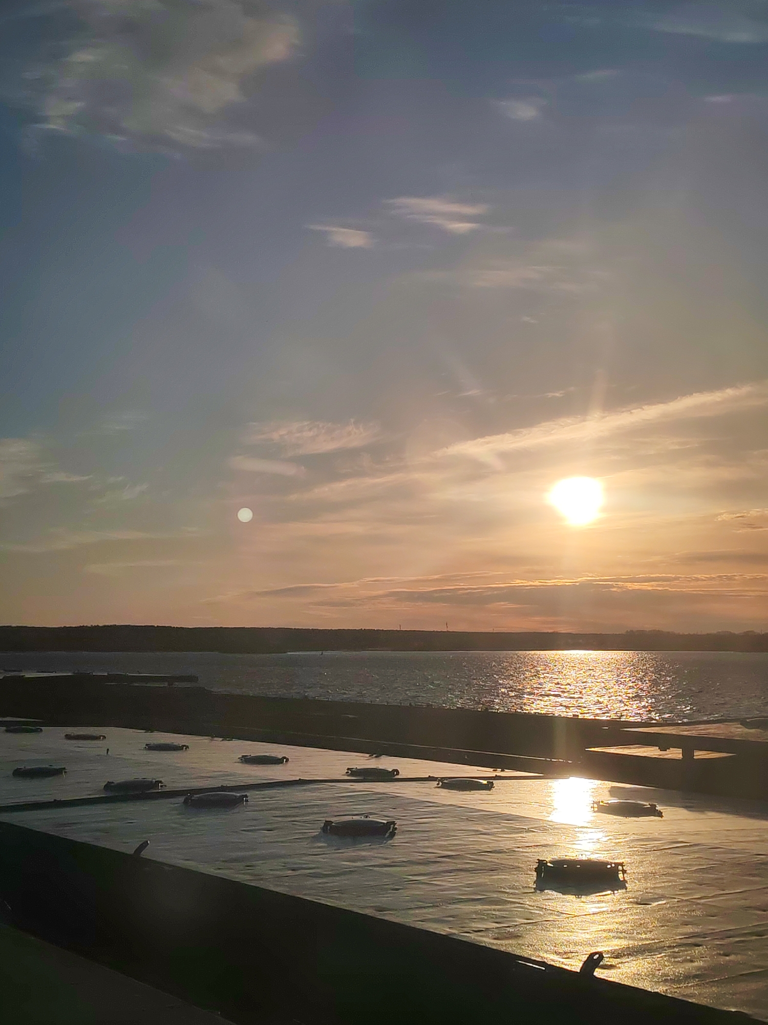 The image captures an expansive coastal or lakeside scene at either sunrise or sunset, bathed in warm, golden light. A brilliant sun dominates the upper right portion of the sky, casting a strong, reflective path of light across a large body of water that occupies the middle ground. The water shimmers intensely, especially where it catches the direct sunlight. A dark, low-lying landmass stretches across the horizon, separating the water from the sky.
The sky exhibits a gradient of colors, transitioning from cooler blue-grey tones in the upper left to vibrant orange and yellow hues closer to the setting or rising sun. Scattered, wispy clouds are visible throughout the sky. A notable detail is a distinct, circular, bright, moon-like object positioned in the upper left quadrant of the sky, appearing opposite the sun.
In the foreground, a dark, flat, man-made structure, resembling a large roof or platform, takes up the lower third of the image. This surface also reflects patches of the golden light. Several dark, oblong objects, appearing to be covered containers or pieces of equipment, are arranged in rows across this foreground structure. The scene evokes a tranquil atmosphere, emphasizing the beauty of natural light over water. No text is visible in the image.
