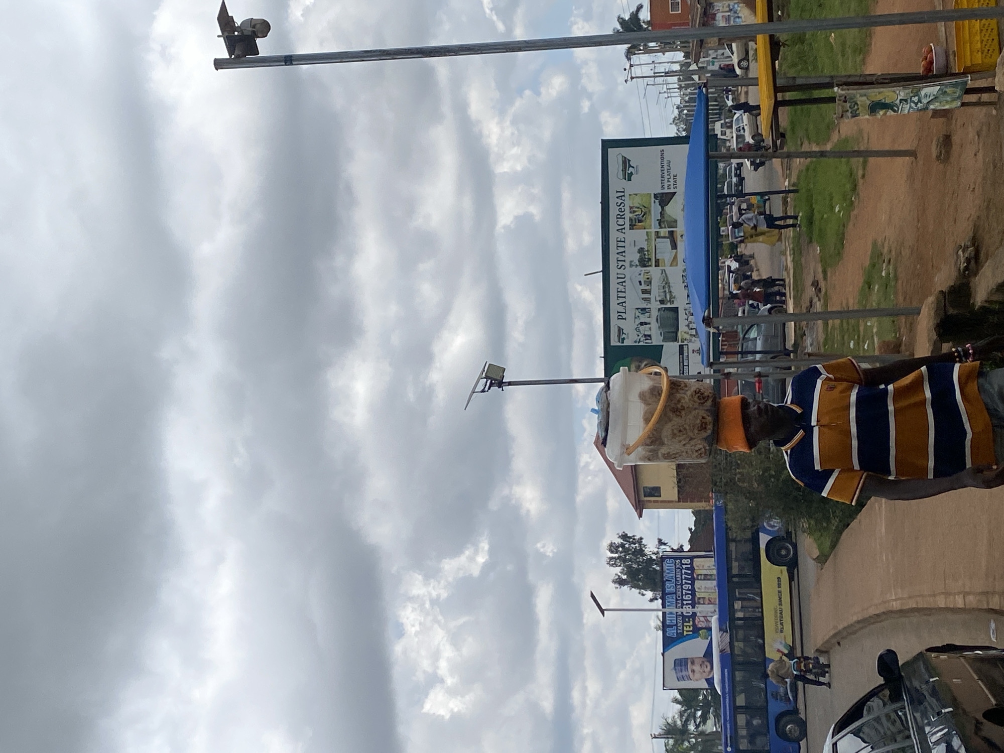 In Bukuru, Nigeria, under a cloudy sky, a man wearing an orange beanie and a striped polo shirt walks along a dirt path, balancing a large bucket filled with goods on his head. The bucket is secured with a yellow strap. To his left, a blue bus with "PLATEAU SINCE 1929" written on its side travels down a road. A billboard behind the bus advertises "AL HIKMA ISLAMIC" with a phone number and a picture of a man. Another larger billboard on the right displays "PLATEAU STATE ACRESAL" with images of various structures and text. In the foreground, a black car is parked on the left side of the road. A blue awning stretches over a market stall area in the middle ground, with several people gathered. Further in the background, more vehicles and buildings are visible. A camera mounted on a pole is positioned in the upper right corner of the frame.