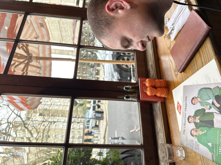 The image depicts an interior view from a window table at "The Orange" restaurant, located in Pimlico, London, United Kingdom.
In the foreground, a wooden table displays a clear glass of water on the left. Adjacent to it, an open copy of "The Observer Magazine" features three individuals on its cover. Behind the magazine, a white paper menu and a brown leather menu holder, embossed with "The Orange" branding, are present. To the right, a set of silver cutlery (fork and knife) rests on a white napkin. Two clear bottles, containing dark and light liquids (presumed oil and vinegar), stand alongside two orange salt and pepper grinders.
On the far right, a male individual with short hair and a small hoop earring in his left ear is partially visible from the chest up, positioned in profile and looking down towards the table.
Through the multi-paned wooden window frame, an orange and white striped patio umbrella is partially seen on the right. The street outside reveals parked vehicles, including a light-colored classic Land Rover Defender and a dark sedan. Two blurry human figures are visible on the sidewalk. Across the street, multi-story brick buildings with numerous windows, typical of London architecture, line the road under an overcast sky.