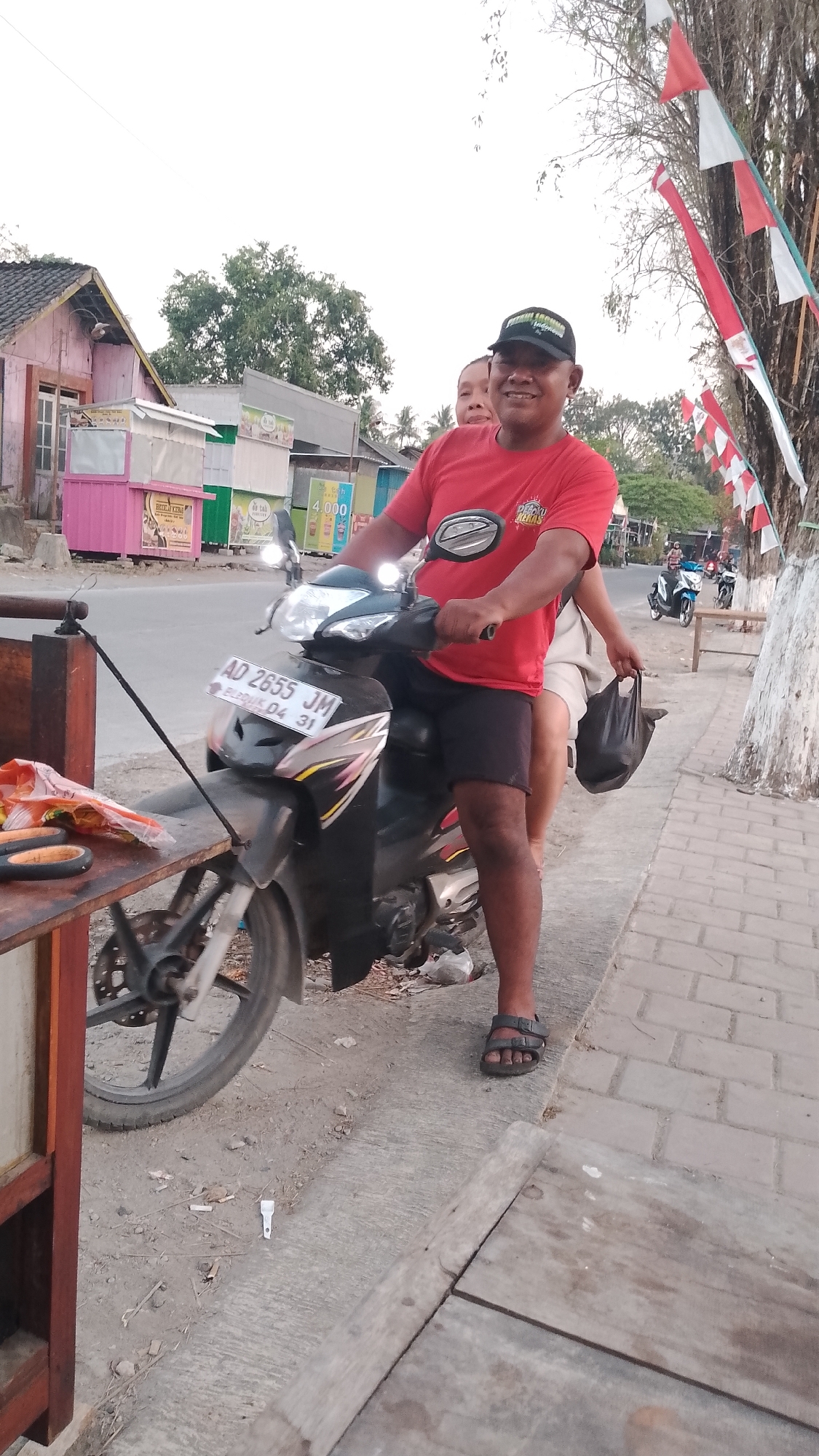 The image depicts two individuals on a motorcycle in what appears to be a street scene in Karanglangu, Indonesia. The person in the foreground, wearing a red t-shirt and dark shorts, is driving the motorcycle. They are smiling and looking towards the camera. Behind them, a second person, partially visible, is seated as a passenger. The motorcycle has a license plate that reads "AD 2655 JM".
The environment suggests a developing or rural area with low-rise buildings lining the street on the left side, some featuring colorful facades and signage, including one that indicates "4.000". A stall or table is set up on the left side of the motorcycle, with orange plastic packaging and what appears to be a pair of scissors on it. A black bag is held by the passenger on the right. Decorative flags, red and white, are visible on poles along the right side of the street. The ground is a mixture of dirt and paved surfaces. The overall lighting suggests it is daytime, possibly late afternoon.
